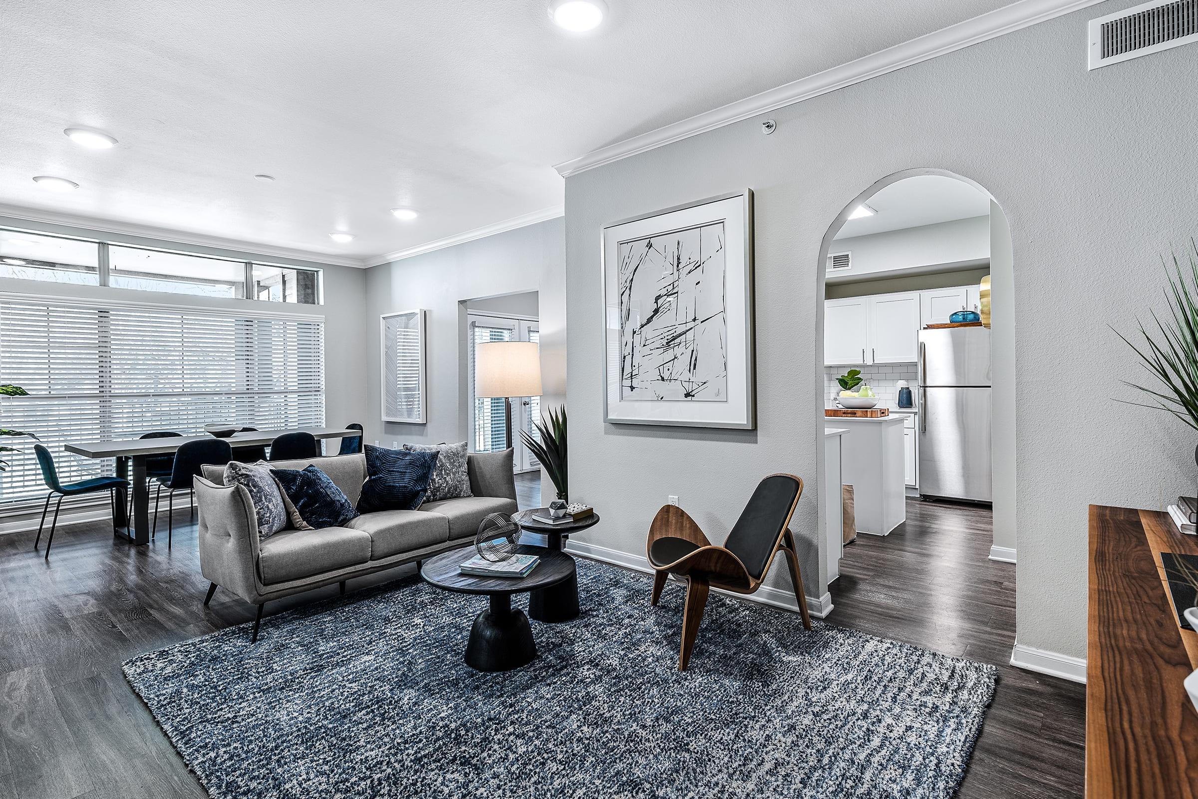 A modern living room with a light gray color scheme, featuring a comfortable gray sofa, a round coffee table, and an accent chair. Large windows provide natural light, and a dining area with a black table and chairs is visible nearby. The decor includes artwork and plants, creating a stylish atmosphere.