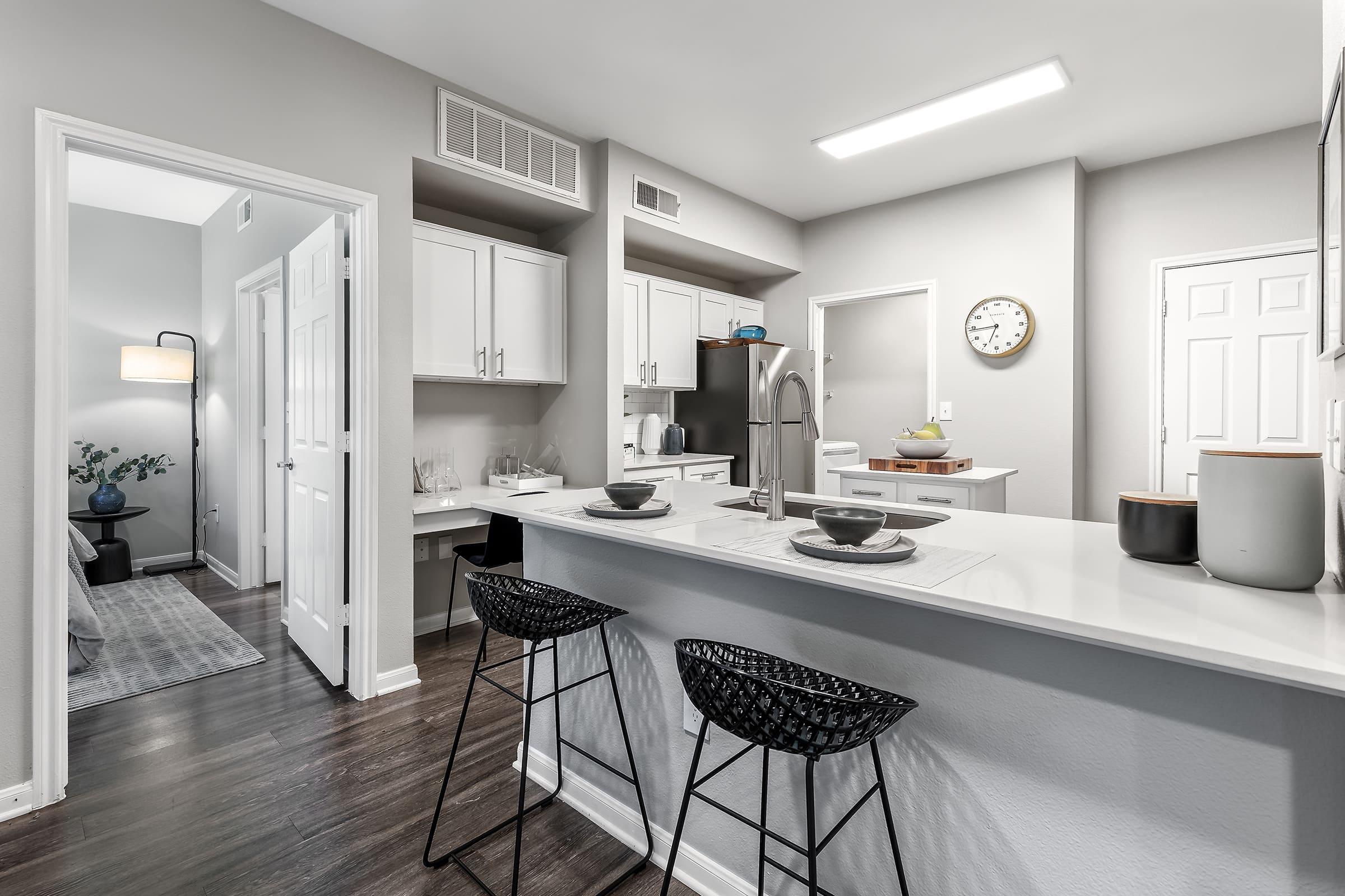 A modern kitchen and dining area featuring a spacious layout with white cabinetry, stainless steel appliances, and a large island with two black bar stools. The room has neutral gray walls and a clock on the wall. A doorway leads to another area, adding an open-concept feel.