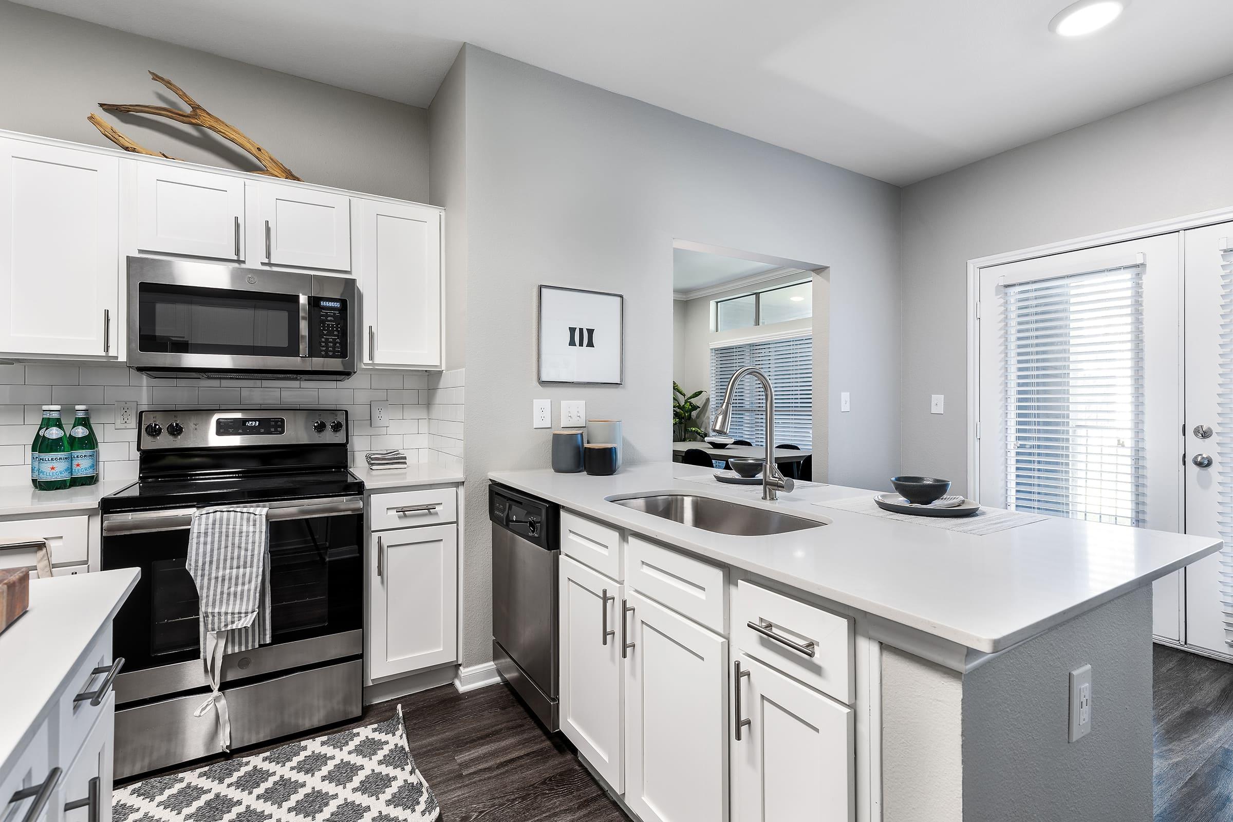 Modern kitchen featuring white cabinetry, stainless steel appliances including a microwave and dishwasher, a double sink, and a stylish countertop. The space is well-lit with natural light and has a neutral color palette, enhanced by decorative elements like a framed wall art and a plant in the background.