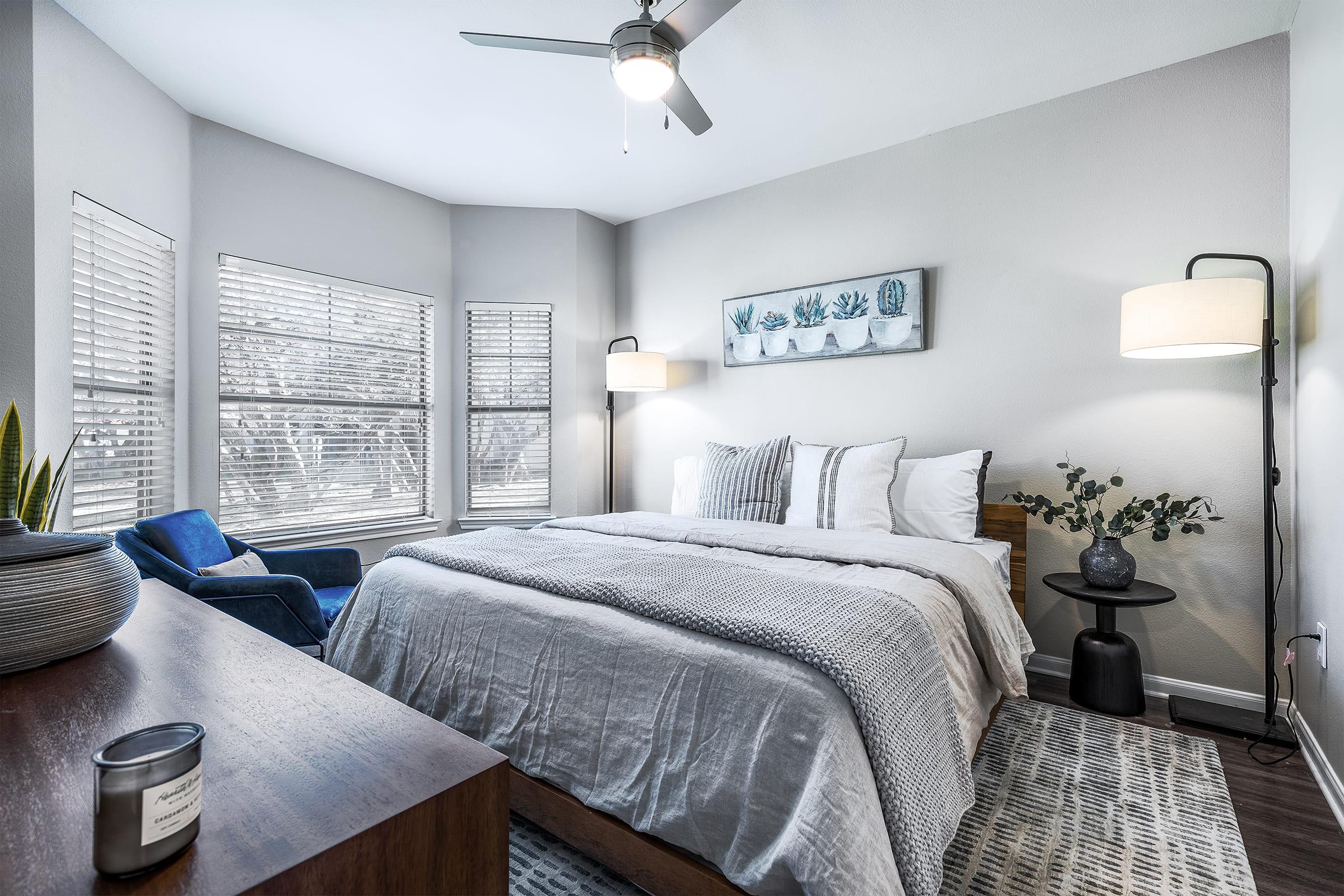 A cozy, modern bedroom featuring a comfortable bed with white linens, decorative pillows, and a light gray blanket. There are two windows with blinds, a blue armchair, a wooden nightstand with a plant and a candle, and a wall art piece showcasing potted plants above the bed. The room is bright and inviting.