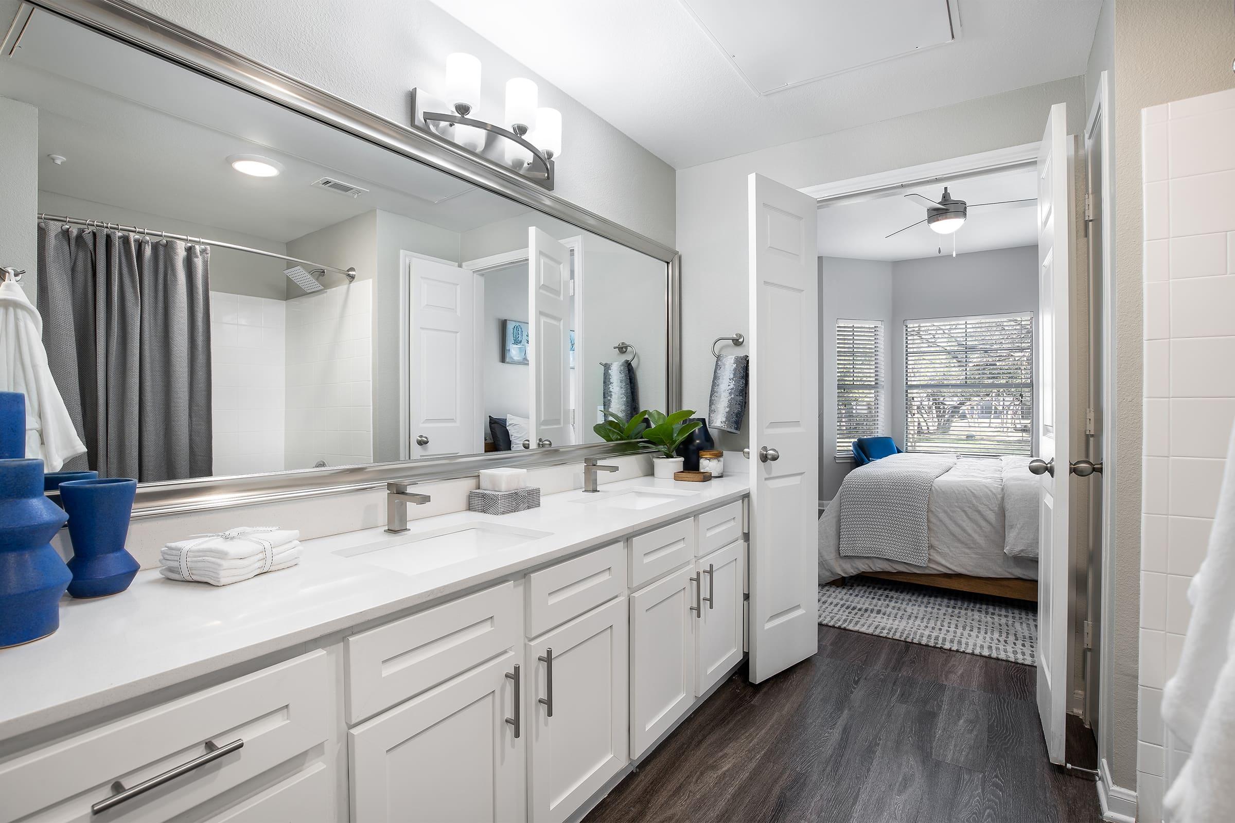 A modern bathroom featuring a double vanity with a large mirror, white cabinetry, and dark wood flooring. There are neatly arranged towels and a plant on the counter. An open door leads to a well-lit bedroom with a ceiling fan and window view of trees outside.