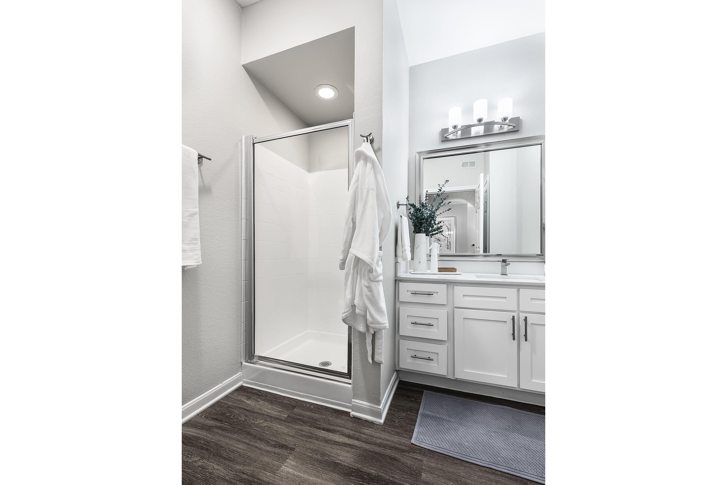 Modern bathroom featuring a glass shower enclosure, a large mirror above a white vanity with silver fixtures, and neatly arranged towels. The warm tones of the wooden floor contrast with the bright white walls and fixtures, creating a clean and inviting atmosphere.