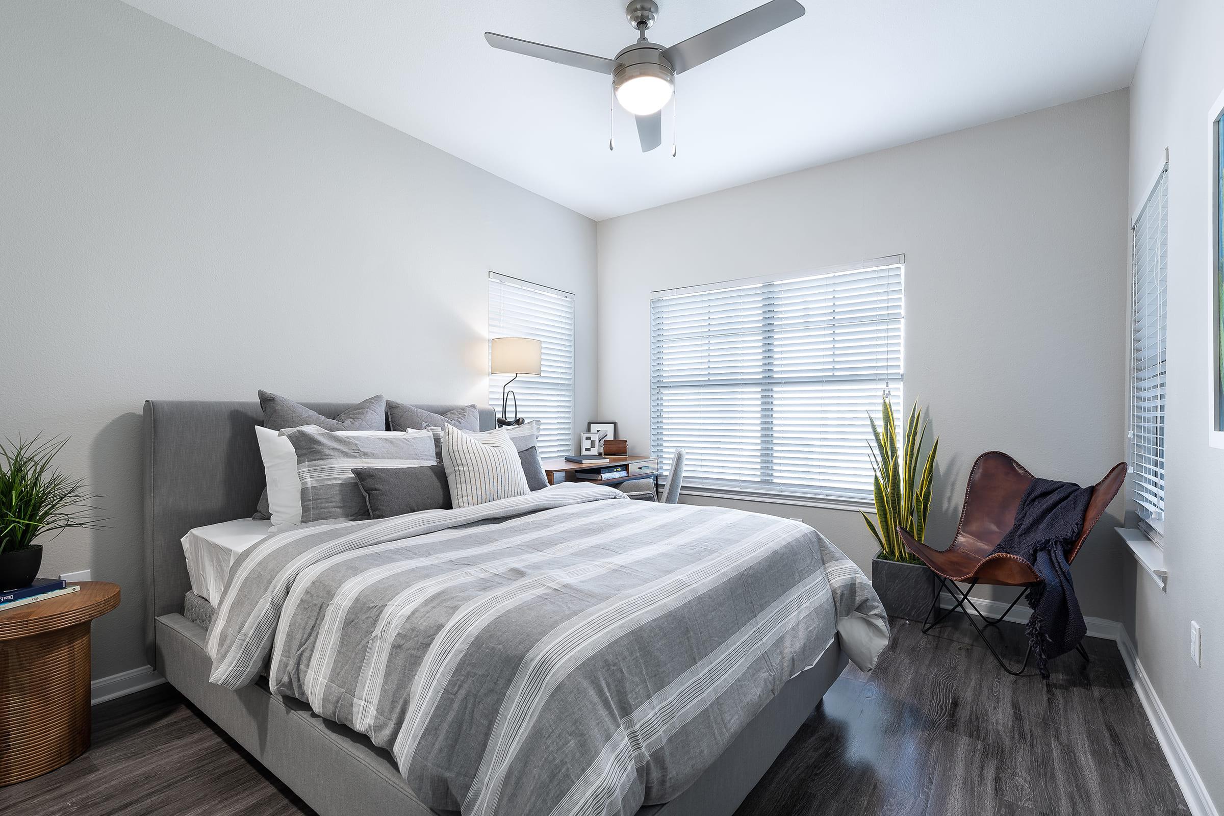 A cozy bedroom featuring a neatly made bed with gray and white striped bedding. There’s a ceiling fan above, a bedside lamp, and two windows with blinds. A small desk is positioned next to the bed, complemented by a potted plant and a stylish leather chair in the corner. The floor is a modern wood finish.