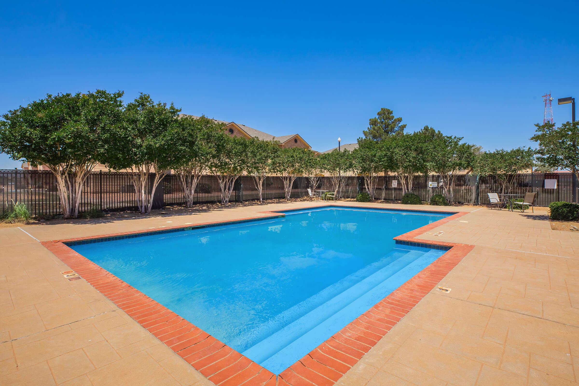 A clear swimming pool surrounded by a brick border in a sunny outdoor space. Lush, green trees line one side of the pool, with a brick patio area. A fence encloses the area, and blue skies are visible above, creating a relaxing atmosphere.
