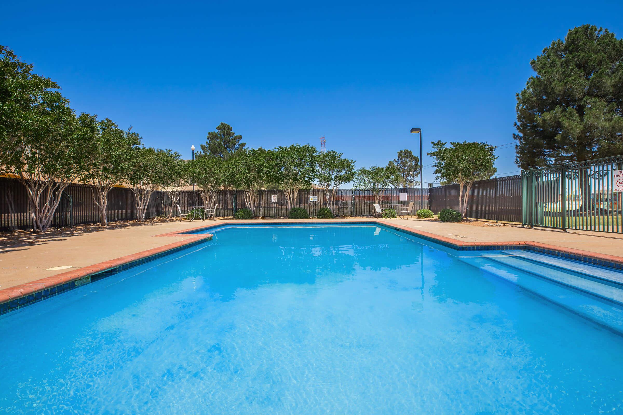 A clear blue swimming pool surrounded by trees and greenery, with a fence in the background. The sky is bright and blue, indicating a sunny day. The pool area features a concrete deck and a few lounge chairs, creating a relaxing outdoor space.