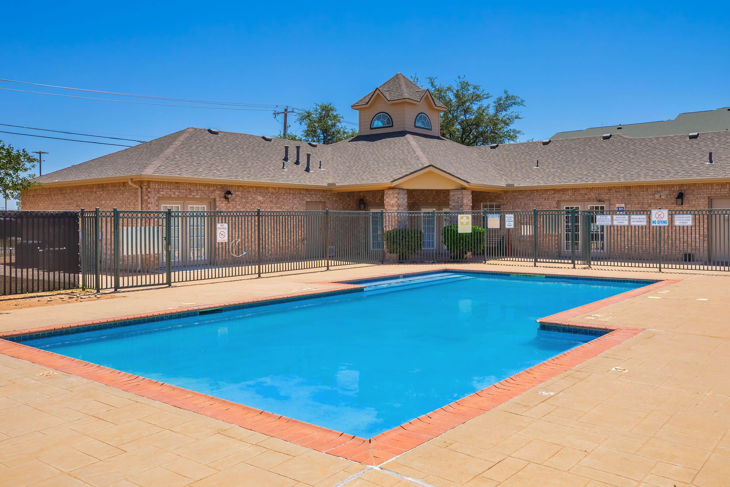 A clear blue swimming pool surrounded by a tan tiled deck. A building with a peaked roof is in the background, featuring multiple windows and a fenced perimeter. Various signs are posted around the pool area, indicating safety and rules. Bright blue sky overhead.