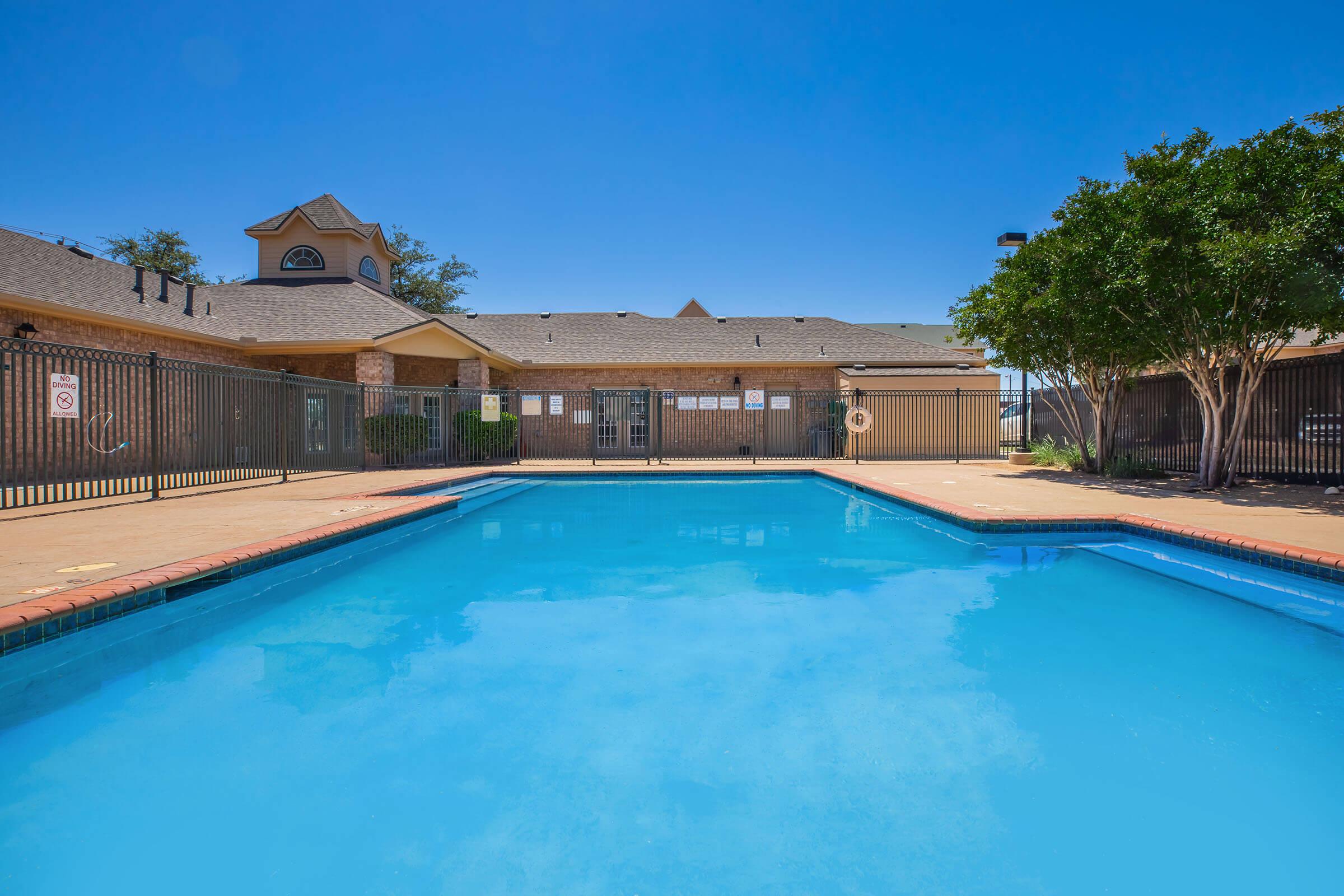 A clear blue swimming pool surrounded by a gated area. The pool is framed by brick pavement, and in the background, there is a tan building with a peaked roof and a few trees. The sky is bright blue with no clouds.