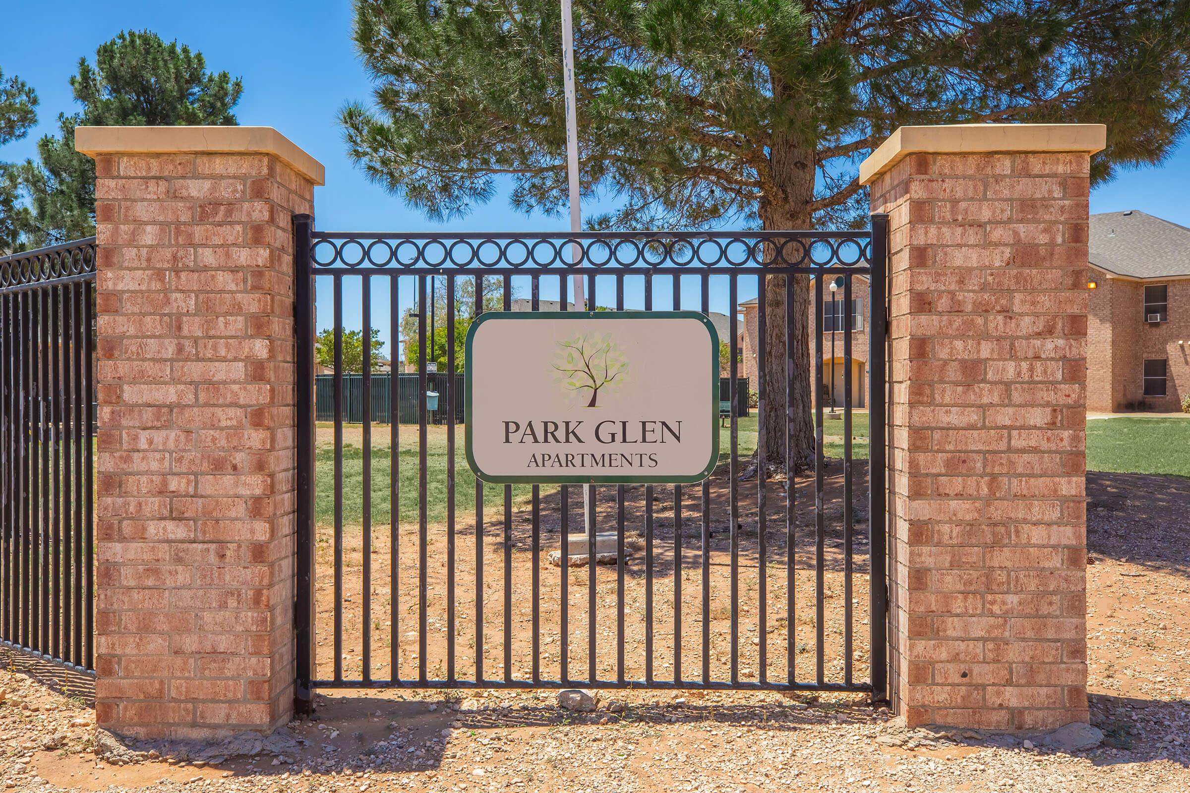 A gated entrance featuring a brick wall and a metal fence. A sign in the center reads "PARK GLEN APARTMENTS," with a tree graphic above. The surrounding area shows grass and trees under a clear blue sky.