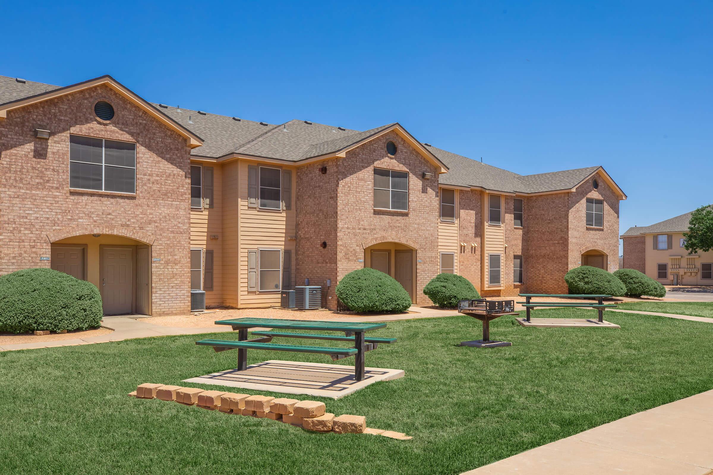 A row of beige and brick apartment buildings with a clear blue sky above. In the foreground, there are well-maintained grassy areas with shrubs and small picnic tables on concrete pads, providing outdoor spaces for residents. The buildings feature large windows and a mix of siding materials.