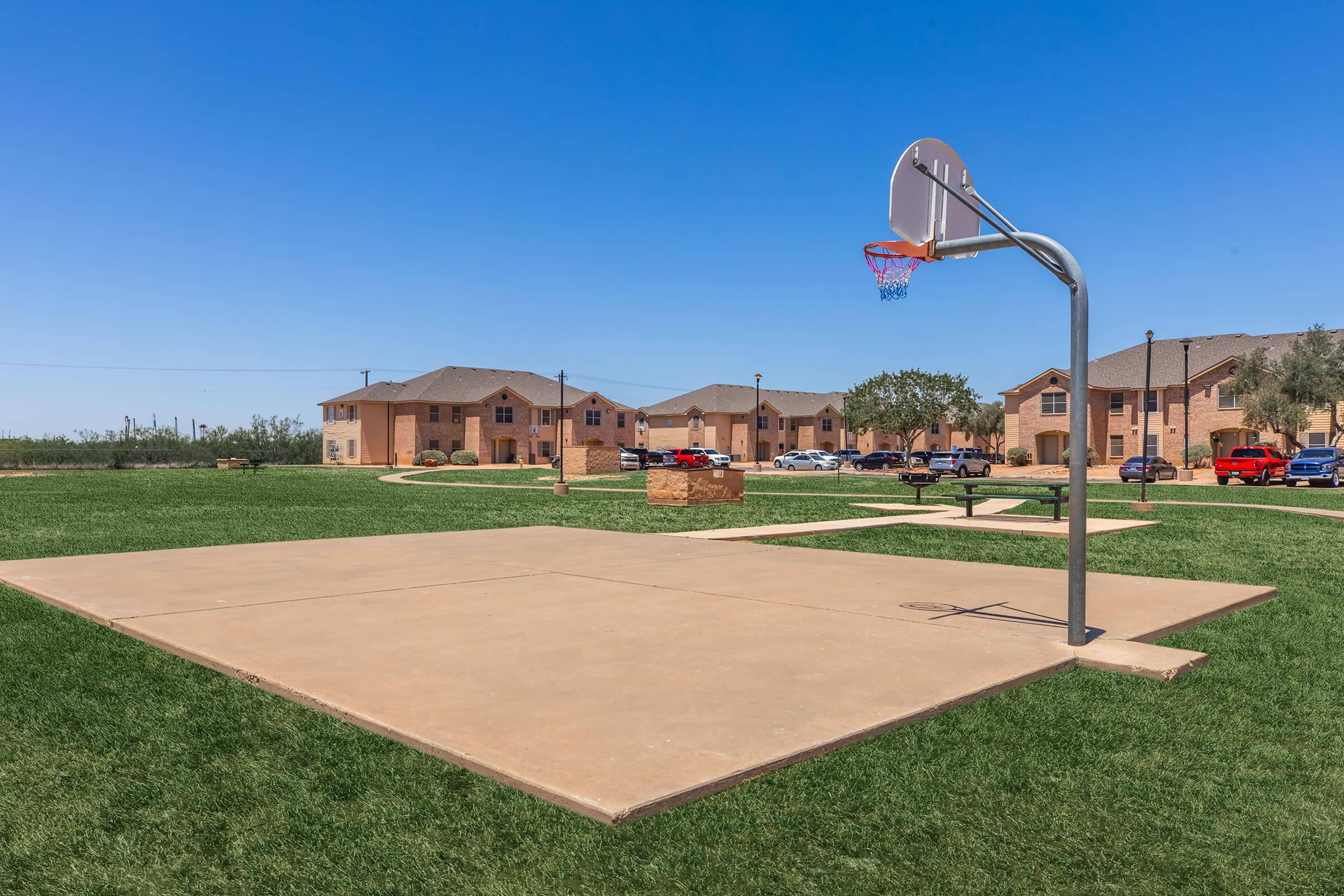 A basketball court with a hoop, situated on a grassy area. In the background, there are several residential buildings and parked cars under a clear blue sky. The scene is bright and sunny, ideal for outdoor activities.