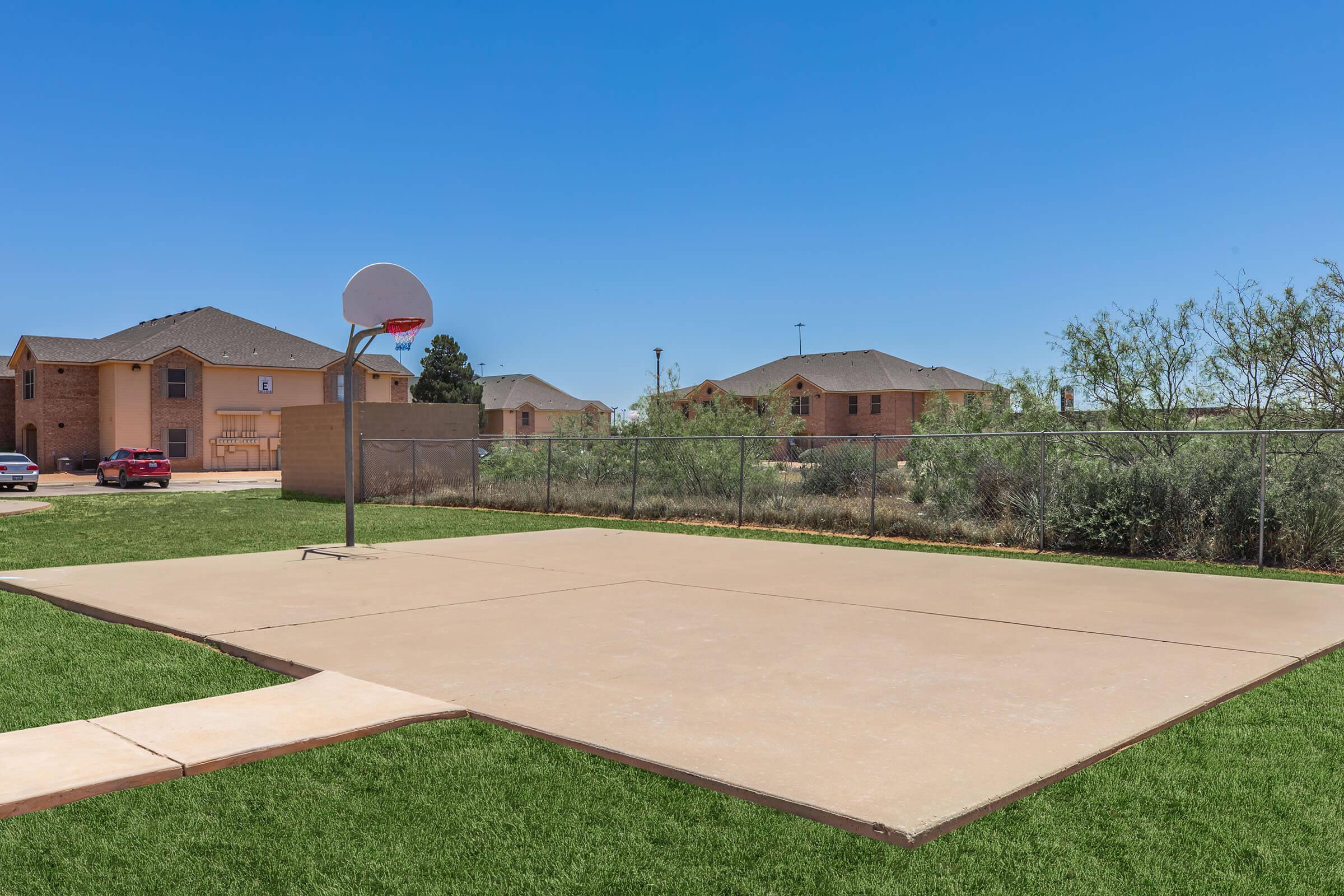 A basketball court with a hoop, located in an outdoor area. The court is surrounded by a fence, with grassy edges. In the background, there are several residential buildings under a clear blue sky.