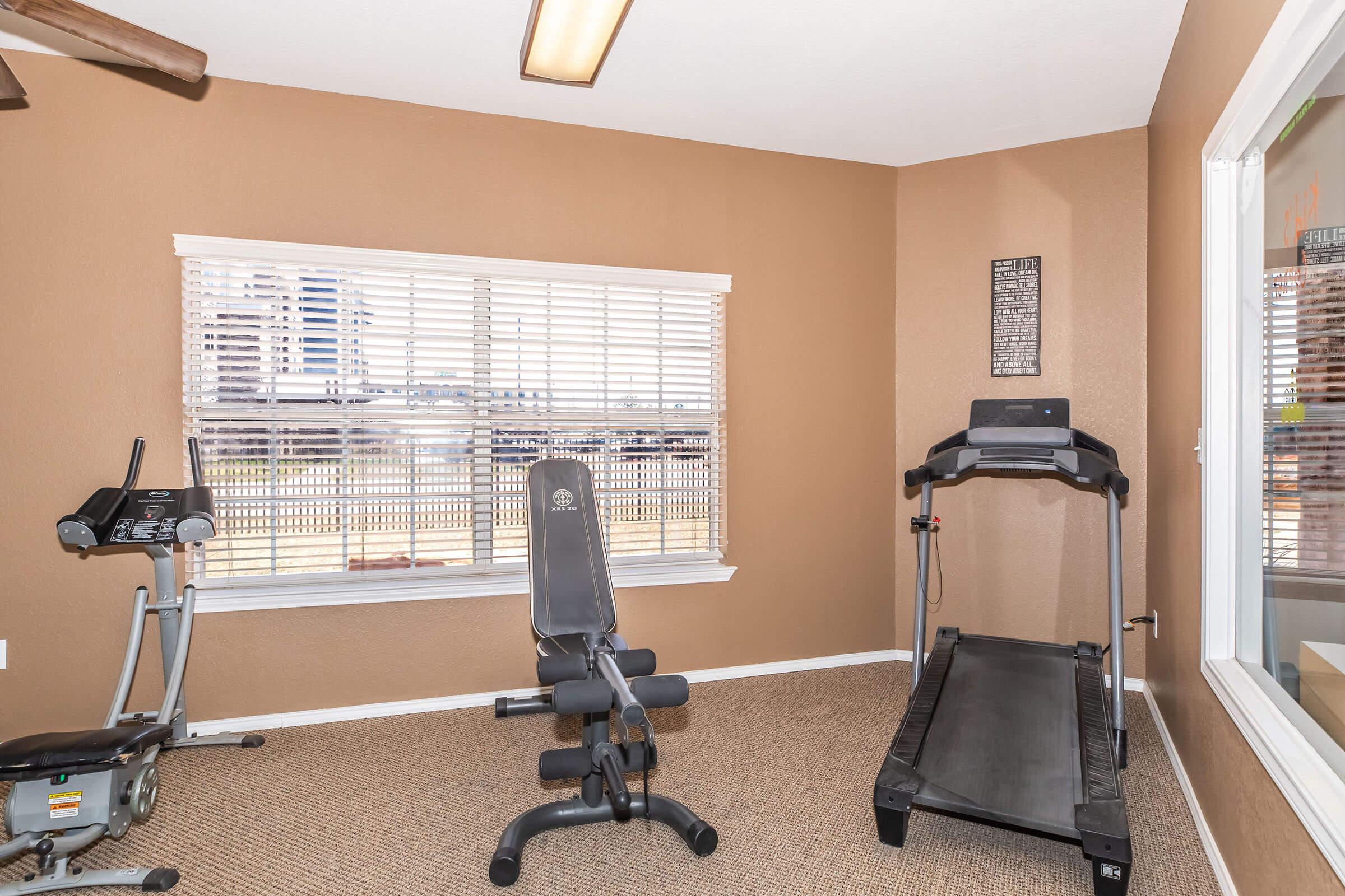 A small home gym with a treadmill, exercise bike, and weight bench. Natural light comes through a window with white blinds, illuminating the room's brown walls and carpeted floor. A motivational poster hangs on the wall, creating a welcoming workout atmosphere.