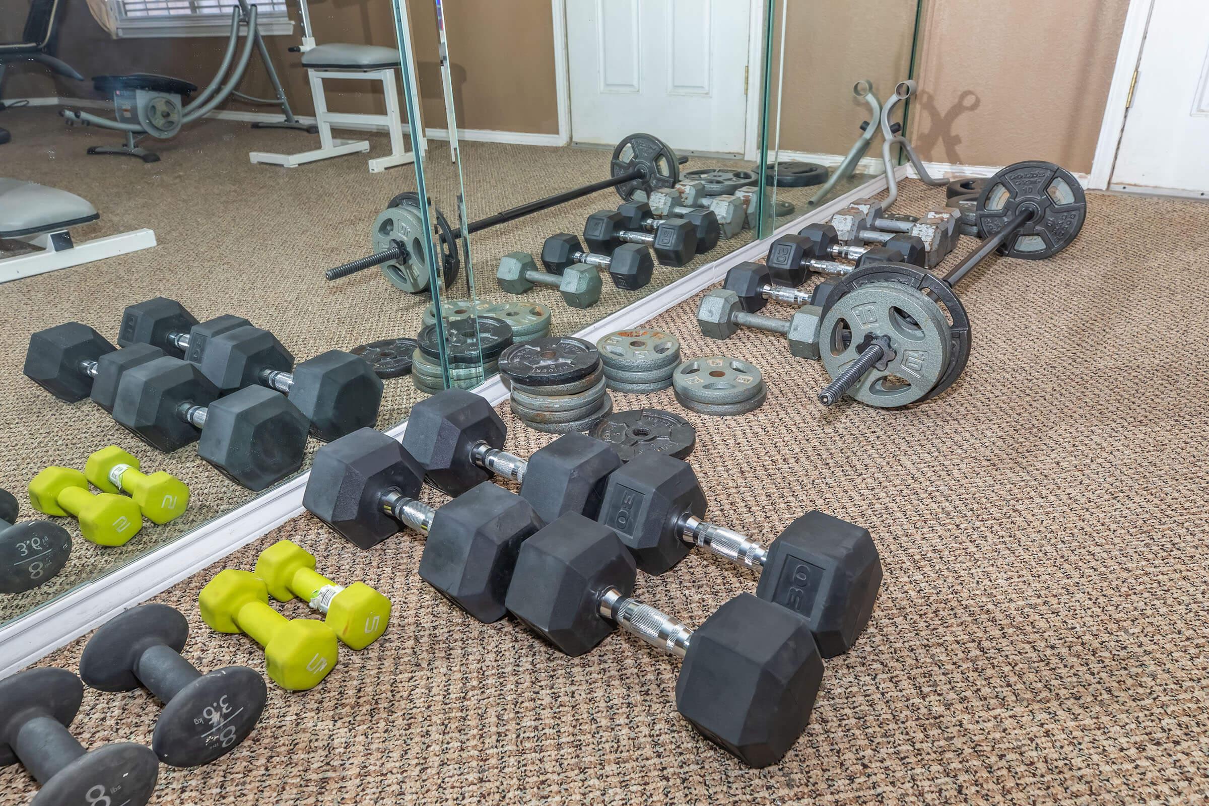 A home gym setting featuring various weights. There are sets of dumbbells in different sizes, including a pair of yellow 5-pound weights, along with a barbell and weight plates on the floor. A mirror reflects the equipment, and the walls are painted a neutral color.