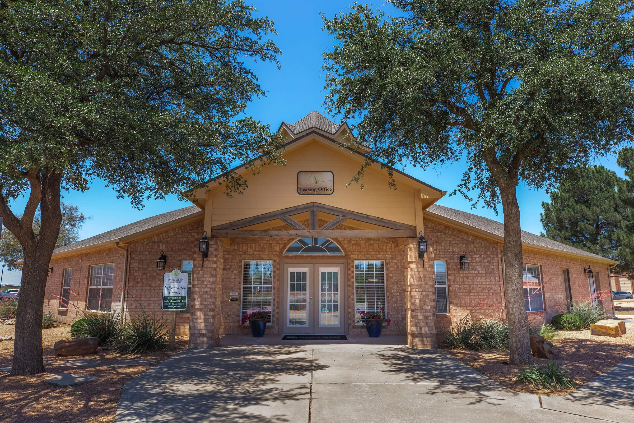 A welcoming single-story building with a light yellow exterior, brick accents, and a triangular roof. Flanked by two trees, the entrance features large glass doors and decorative windows. Potted flowers are placed on either side of the entrance, with a clear blue sky overhead.