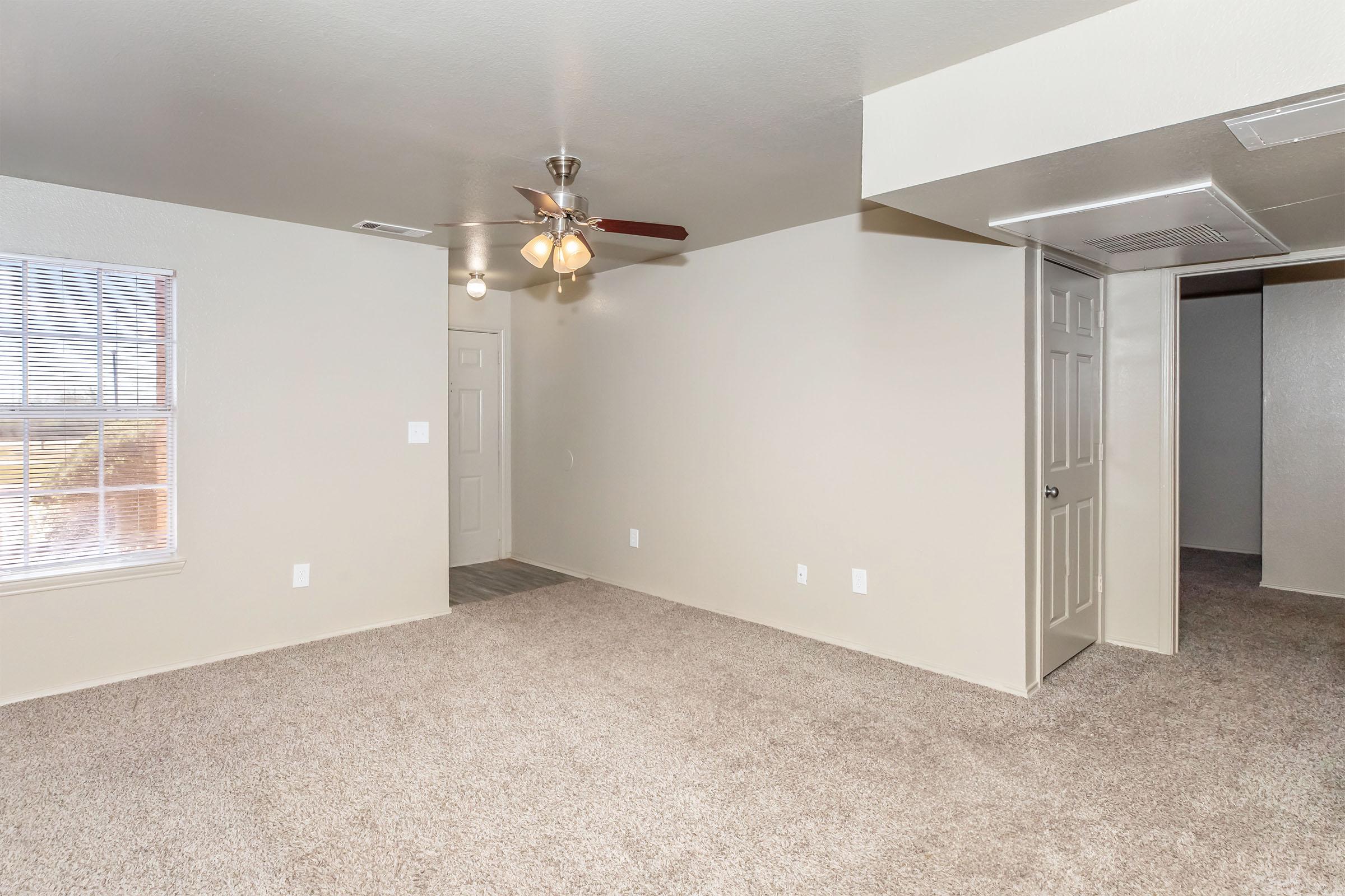 A spacious, empty living room featuring light beige walls and plush carpet. A ceiling fan with light fixtures is installed, and there are windows allowing natural light. A door leads to another room, and the space offers a clean, modern look, ready for furnishing.