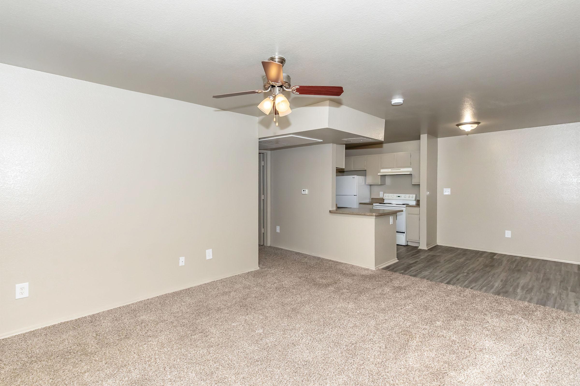 Interior view of a spacious living area with light beige walls and plush carpet. A ceiling fan with light fixture hangs above, and to the left, an open doorway leads to a kitchen area with white appliances and cabinets. The layout is simple, creating a bright and inviting atmosphere.