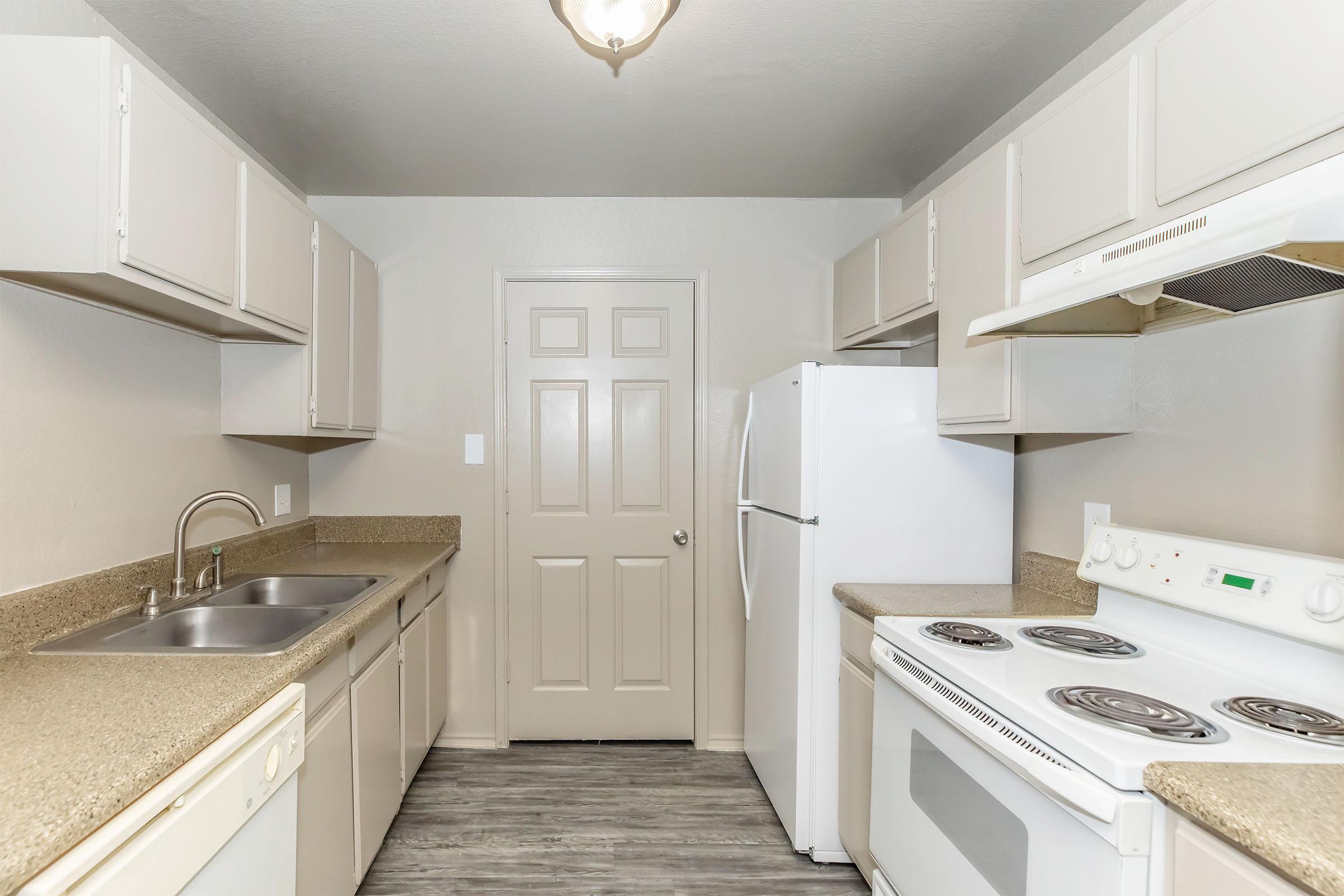 Modern kitchen featuring beige cabinets, a light gray wall, and a door leading to another room. Includes a double sink, white refrigerator, and a stove with an oven. The countertops are a matching beige, and the flooring is a light wood-like material. Ceiling light fixture provides illumination.
