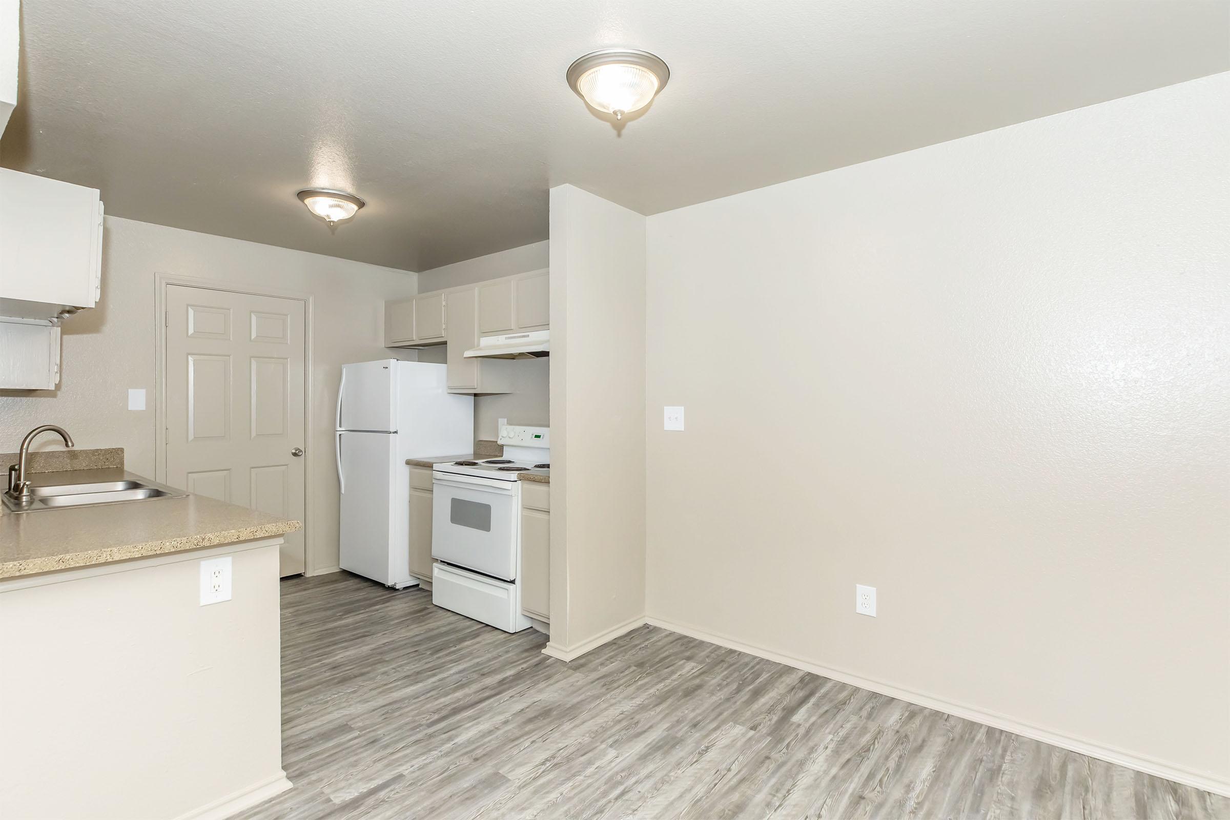 A modern kitchen and dining area featuring light-colored walls, a refrigerator, an oven, and a countertop. The space has laminate flooring and is well-lit by overhead fixtures. A door leads to another room, creating an open and inviting atmosphere.