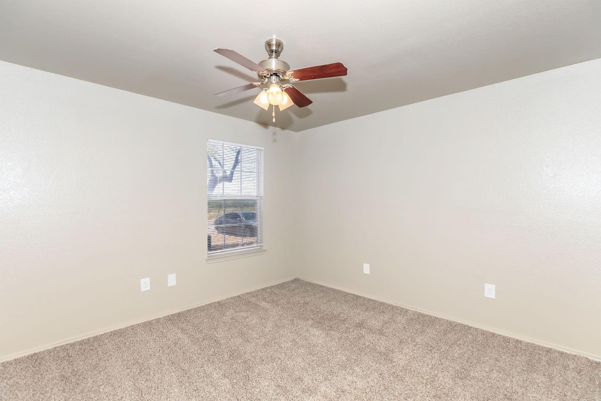 A well-lit, empty room featuring neutral-colored walls and carpet. There is a ceiling fan with three lights, and a window allows natural light to enter. The view outside includes trees. The space is spacious and ready for personal furnishings.