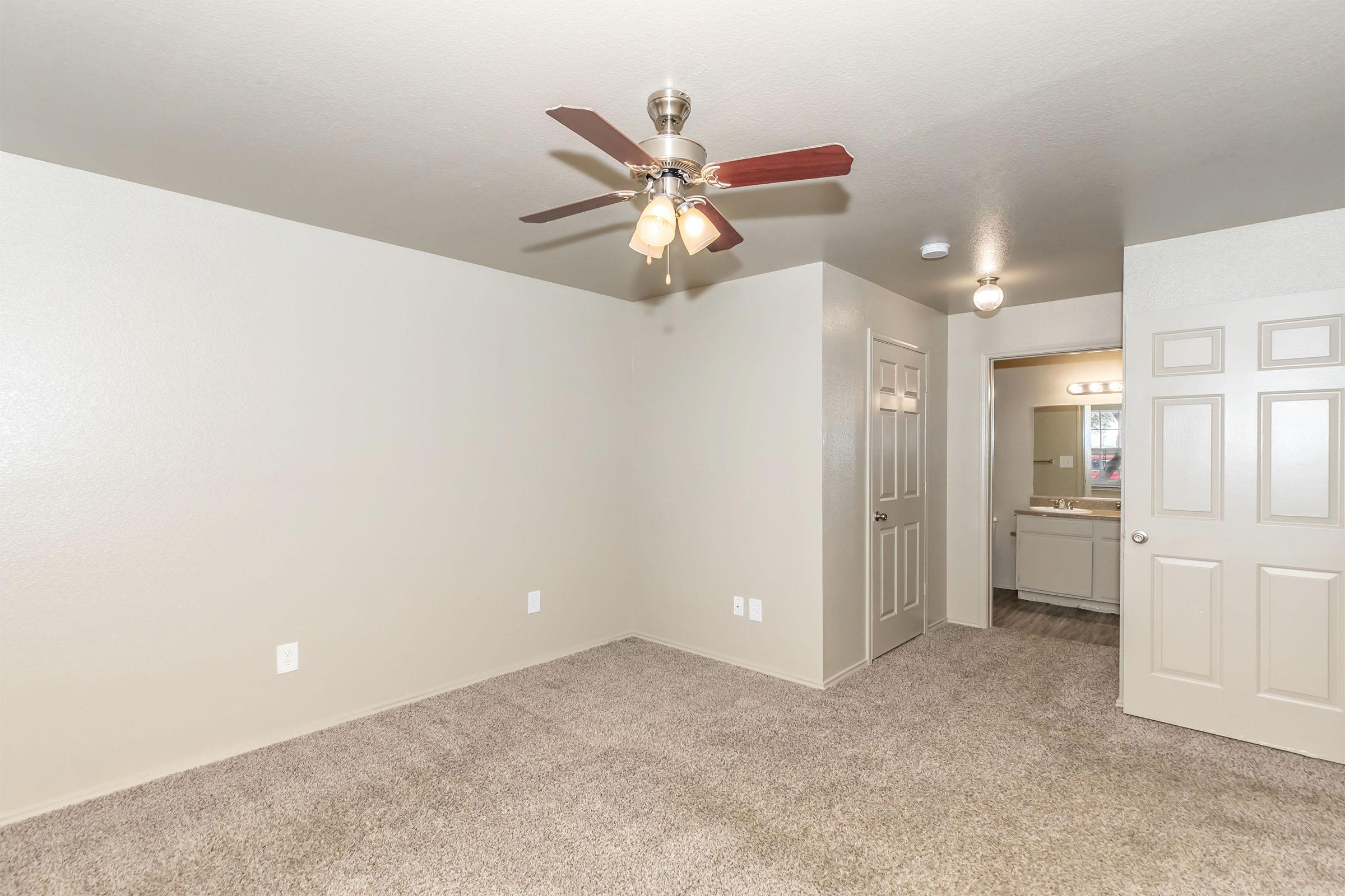 A spacious room featuring light-colored walls and carpet flooring. The ceiling has a ceiling fan with three light bulbs. There are two doors leading to other spaces, and a bathroom area is visible in the background with vanity lights. The room is well-lit and inviting, perfect for various uses.