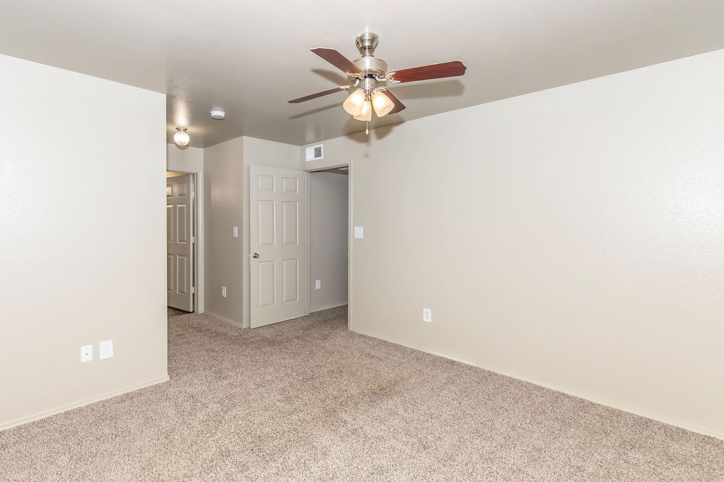 A spacious, empty room with beige walls and light brown carpet. A ceiling fan with four light fixtures hangs from the ceiling. One door leads to another room, and two light fixtures provide additional illumination. The overall atmosphere is bright and clean, ready for furniture and decoration.