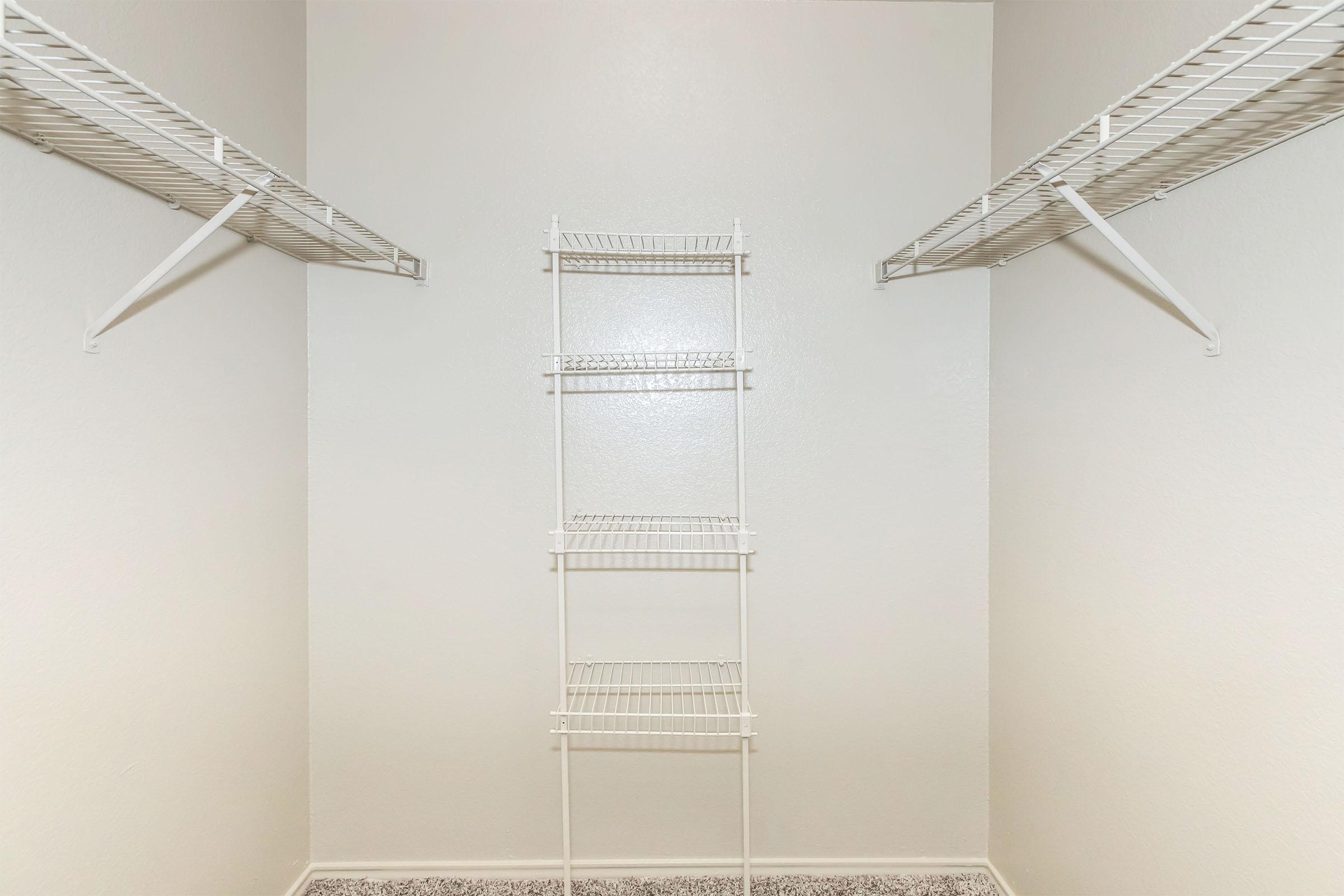 An empty closet with white wire shelves on the sides and a tall wire shelving unit in the center, all set against a light-colored wall. The floor is covered with a soft carpet, creating a clean and uncluttered look.