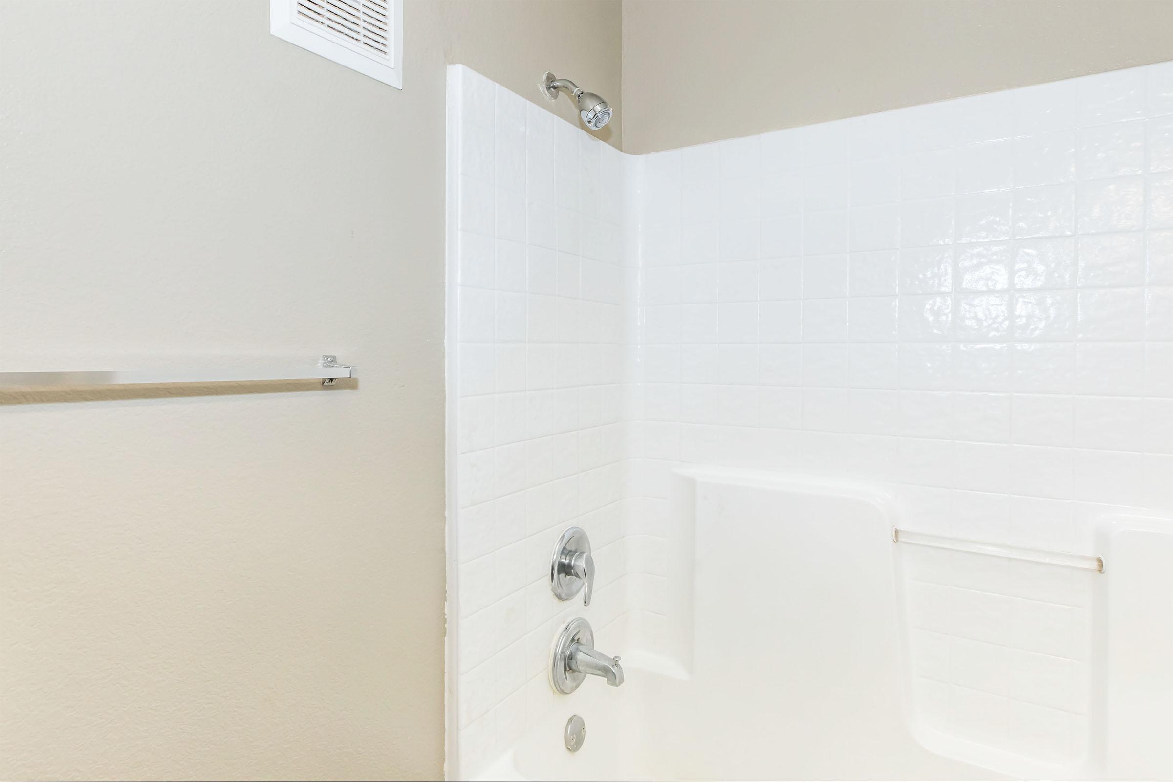 A clean, modern bathtub and shower area with white tiled walls. There’s a chrome showerhead attached to the wall and a tub spout below. A towel bar is visible on the wall beside the shower. The overall color scheme is neutral and bright, creating a fresh and spacious atmosphere.