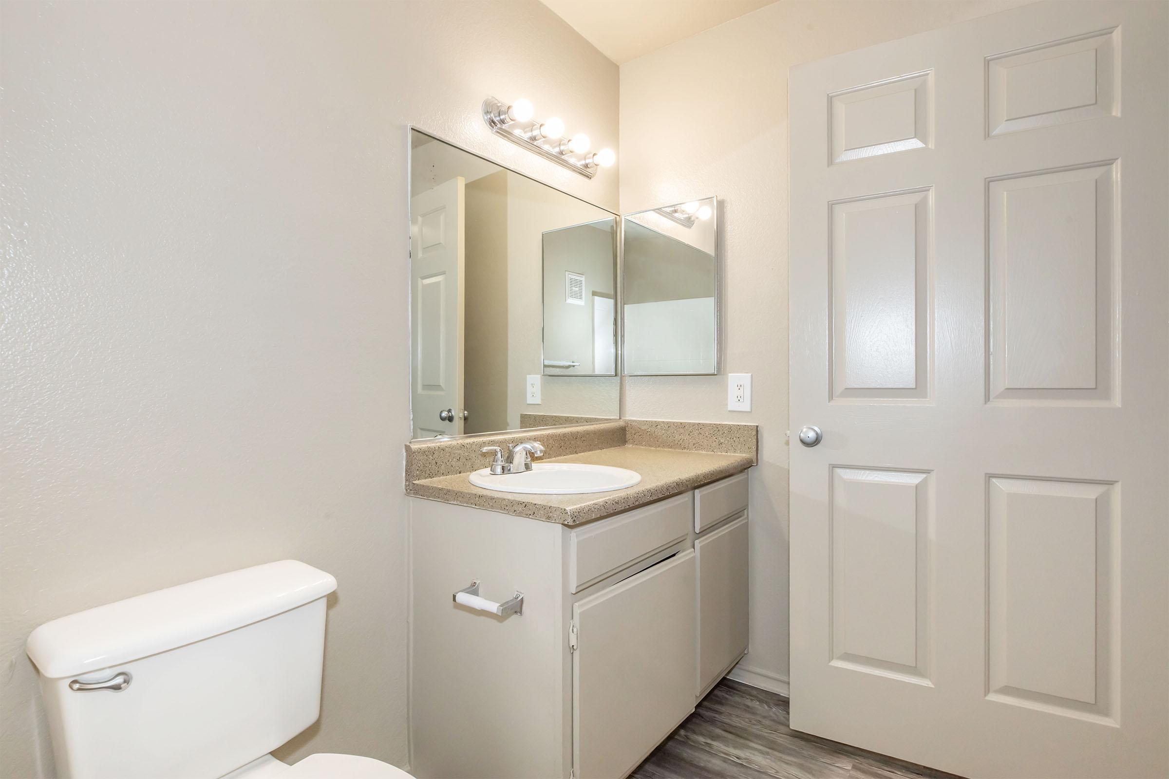 A simple bathroom featuring a light-colored wall and a large mirror above a countertop with a sink. The space includes a white toilet and a door leading to another room. Bright lighting is provided by a fixture above the mirror. The flooring appears to be a light wood or laminate.