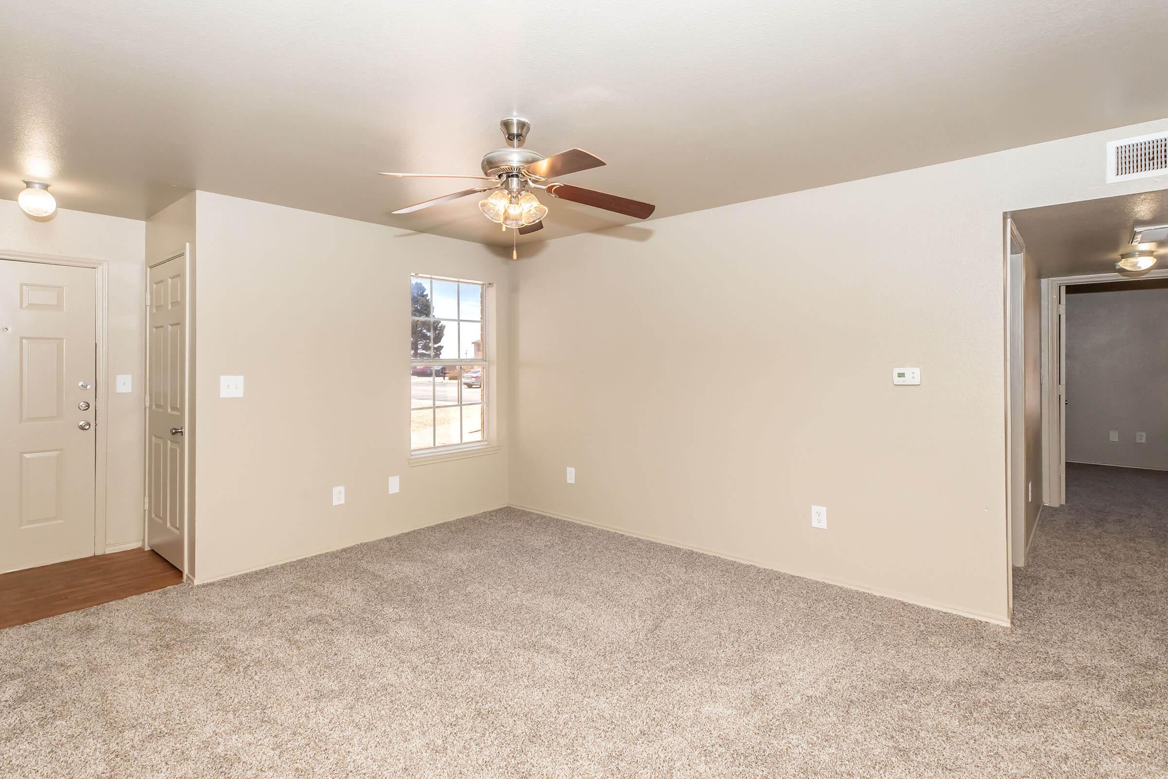 A spacious living area with beige walls and carpeted flooring. It features a ceiling fan and a window allowing natural light. A door leads to an entrance, and another doorway on the right suggests access to additional rooms. The overall ambiance is inviting and well-lit.