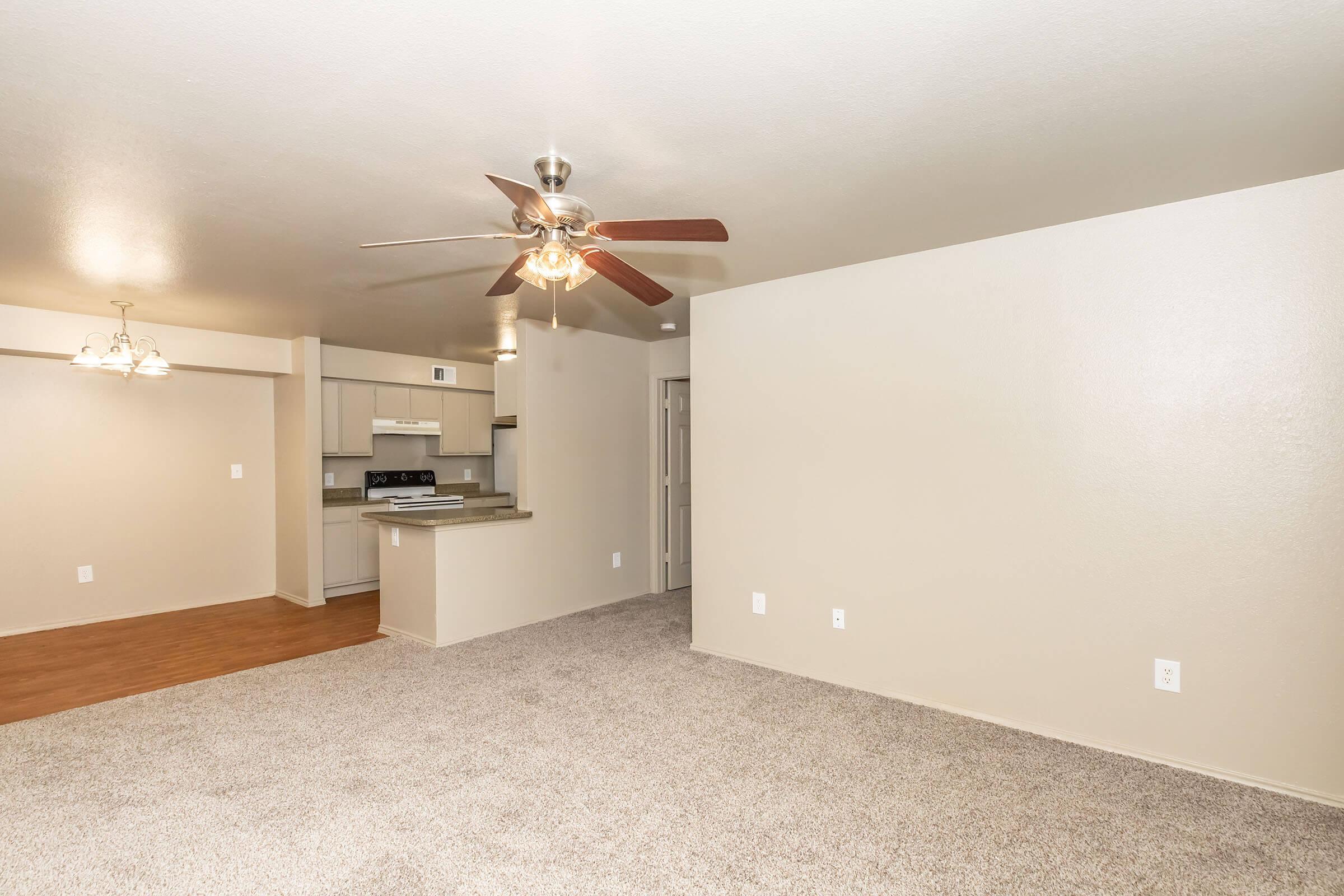 Spacious living area with light beige walls and carpet, featuring a ceiling fan. On the left, a small kitchen area with modern appliances is visible, and a doorway leads to another room. The overall ambiance is bright and inviting, suitable for a cozy home setting.