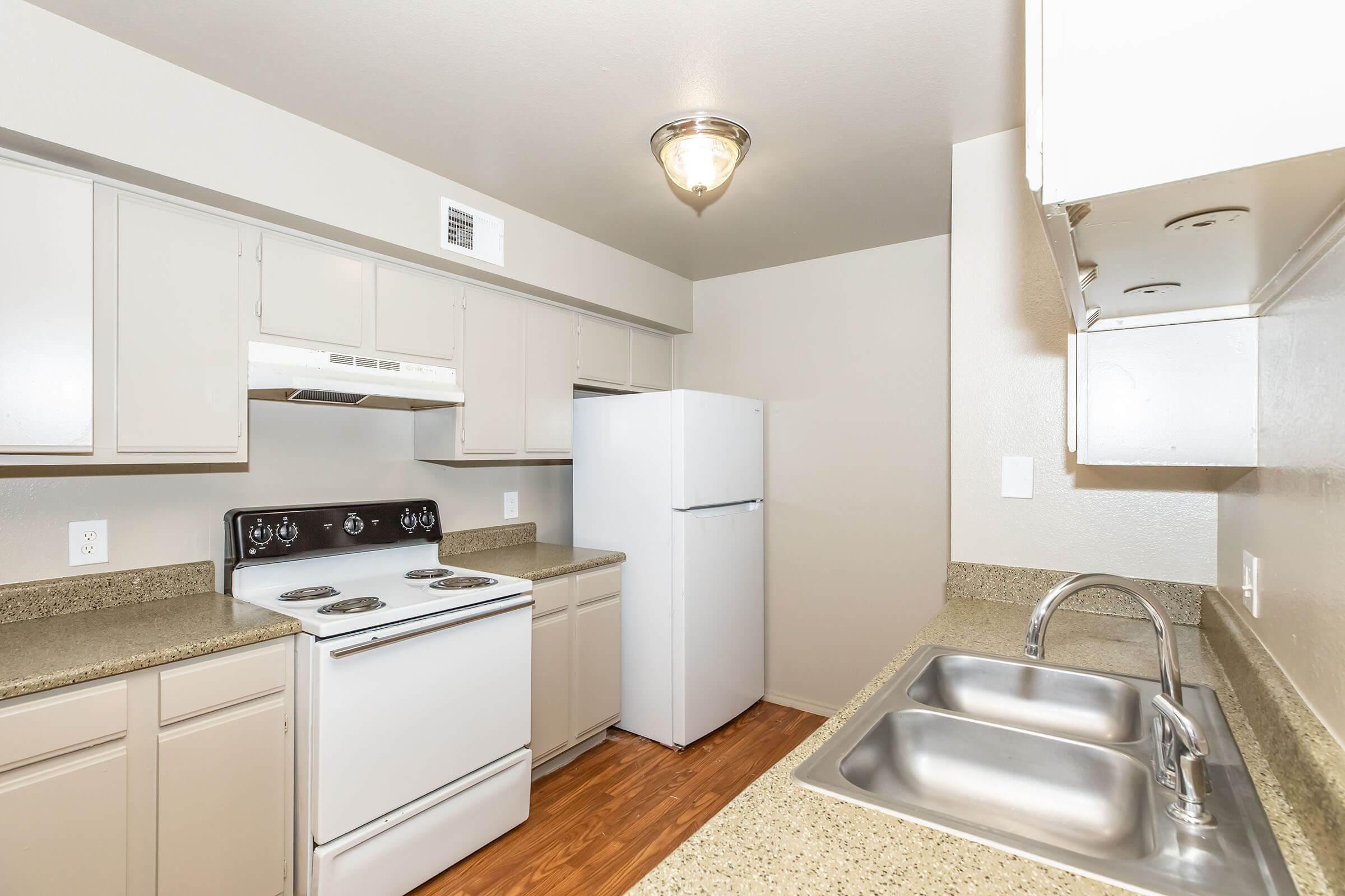 A modern kitchen featuring light-colored cabinets, a white refrigerator, a white stove with an overhead microwave, and dual stainless steel sinks. The countertops are a speckled light brown, and the flooring is a warm wood tone. The space is well-lit with a ceiling fixture.