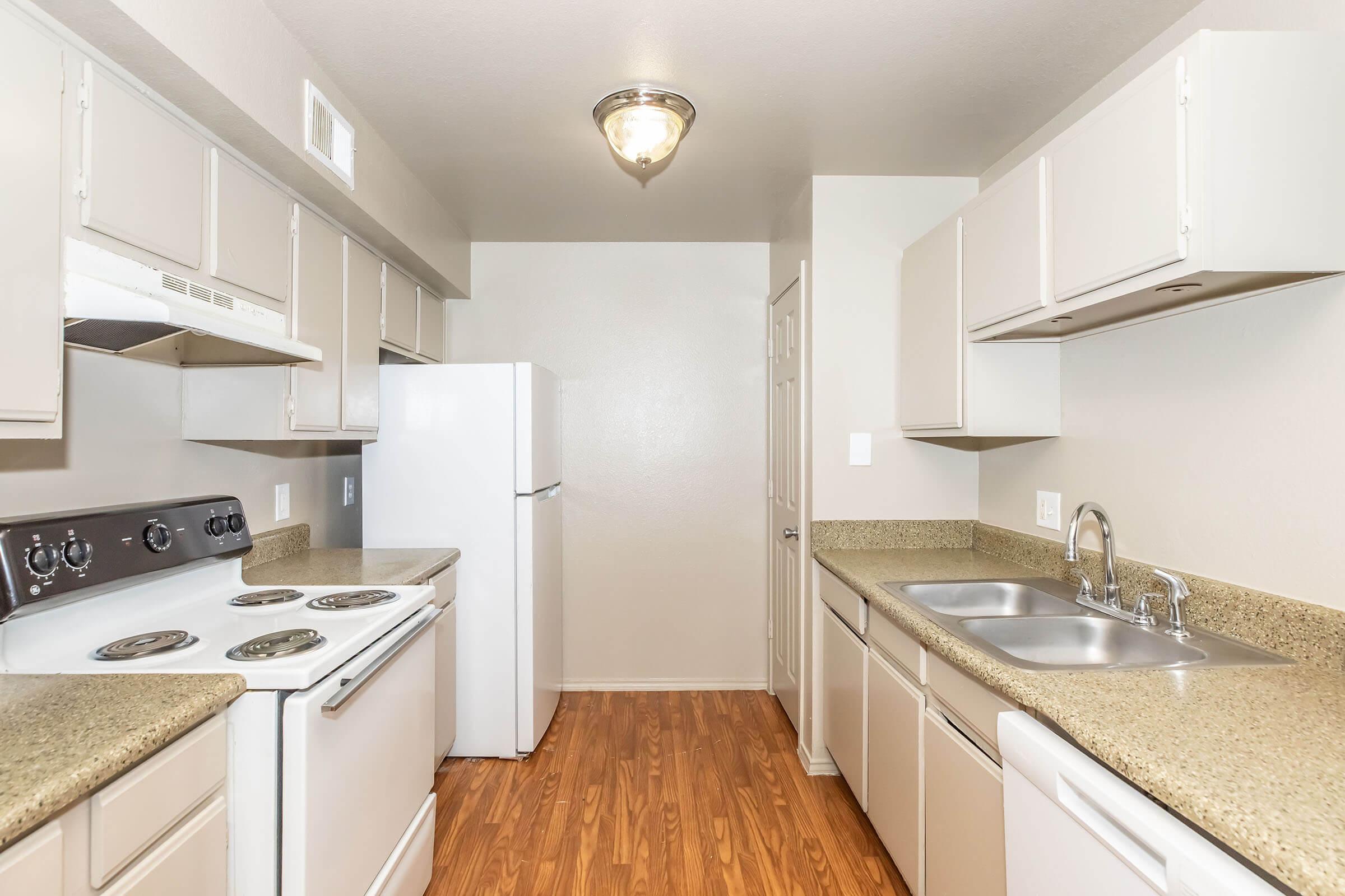 A modern kitchen featuring light-colored cabinets, a white refrigerator, an electric stove, and a double sink. The countertops are a speckled tan material, and the flooring is a warm, laminate wood. The walls are painted a light gray, providing a bright and open feel to the space.