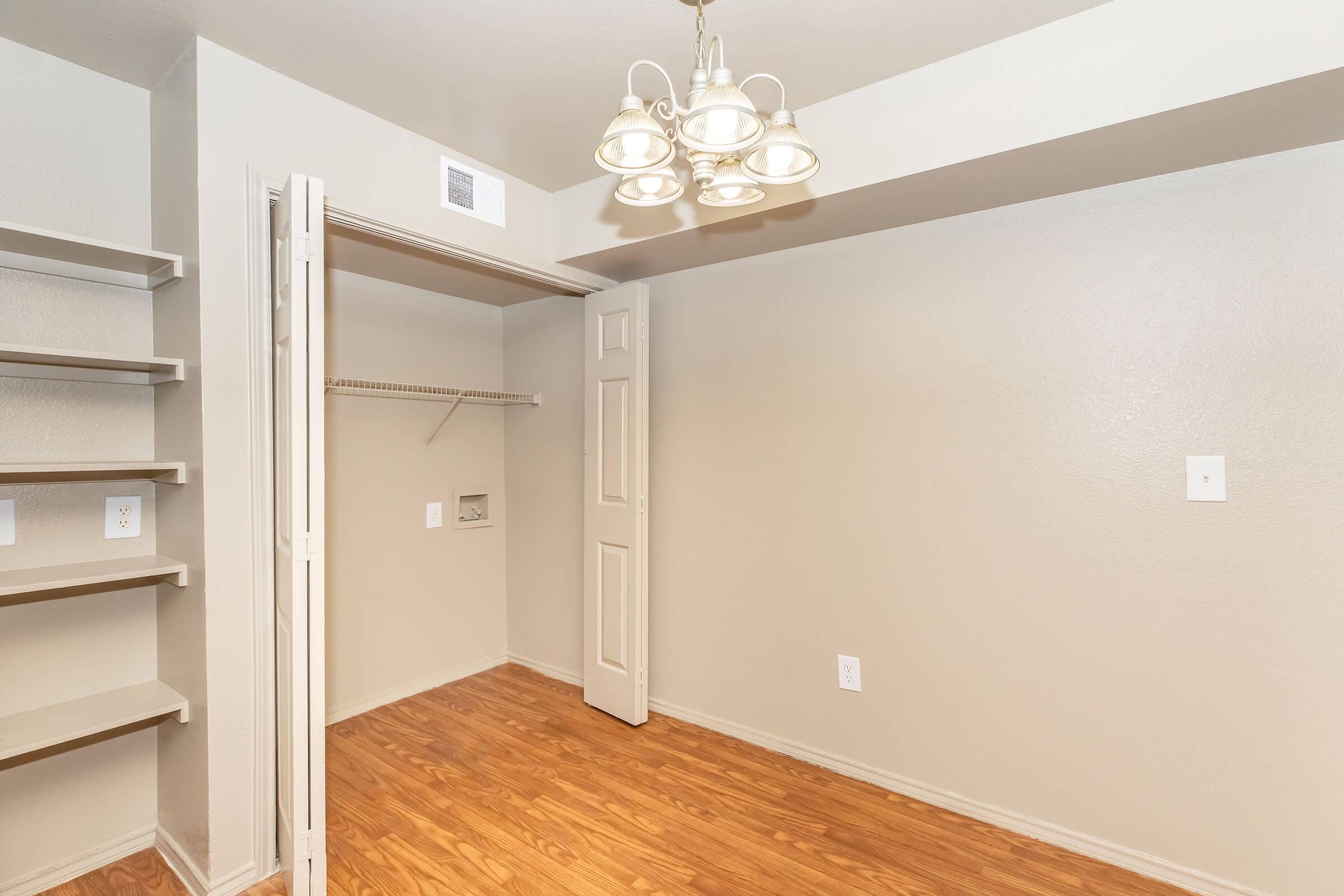 A well-lit room with light beige walls and wooden laminate flooring. There is a closet with double doors on one side and a hanging shelf above. A ceiling light fixture with multiple bulbs provides illumination. No furniture is present, creating an open and spacious feel.