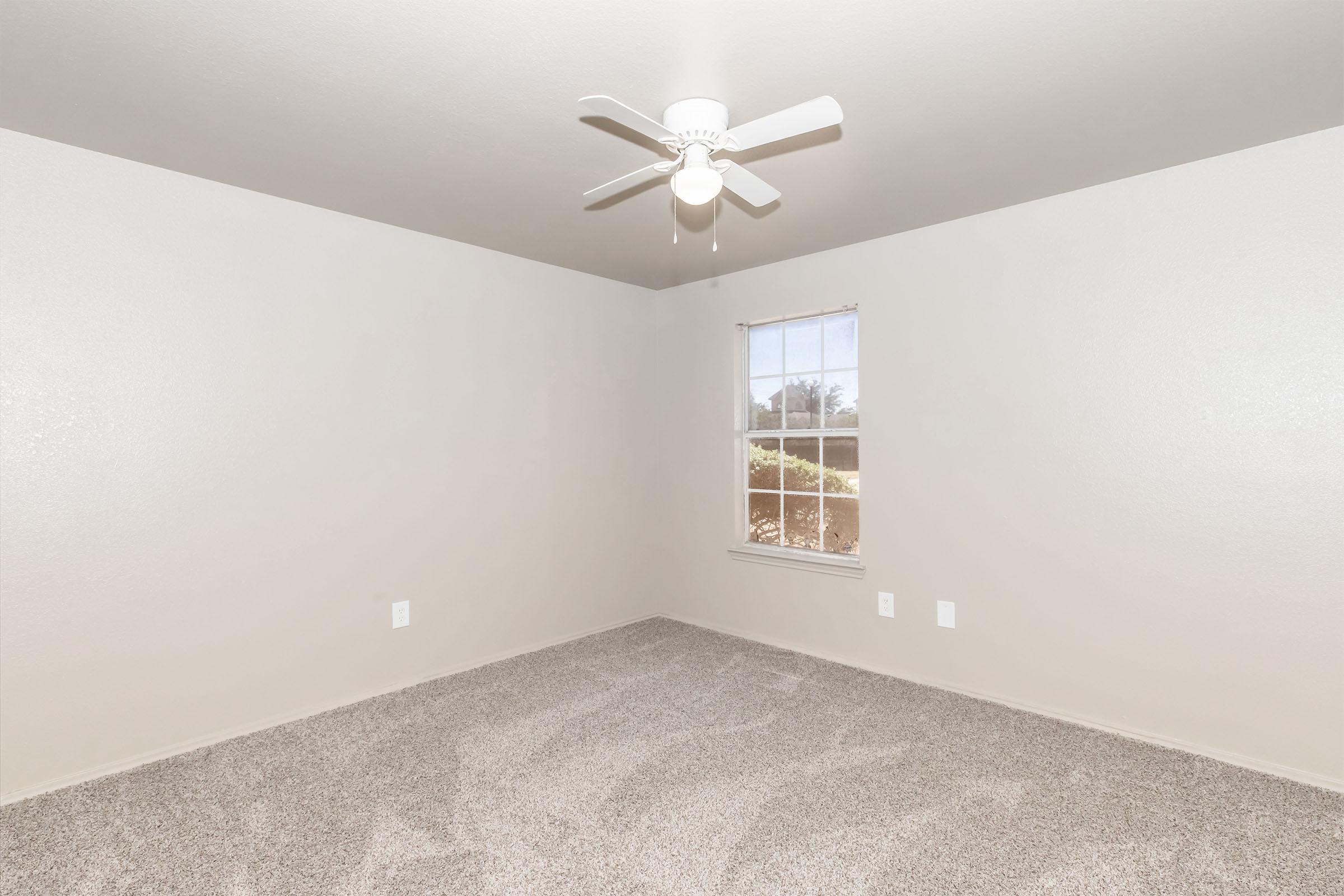 Empty room with light gray walls and carpet. A ceiling fan with three blades is mounted in the center, and there's a window on one wall allowing natural light to enter. The window has a view of greenery outside, adding a touch of nature to the space.