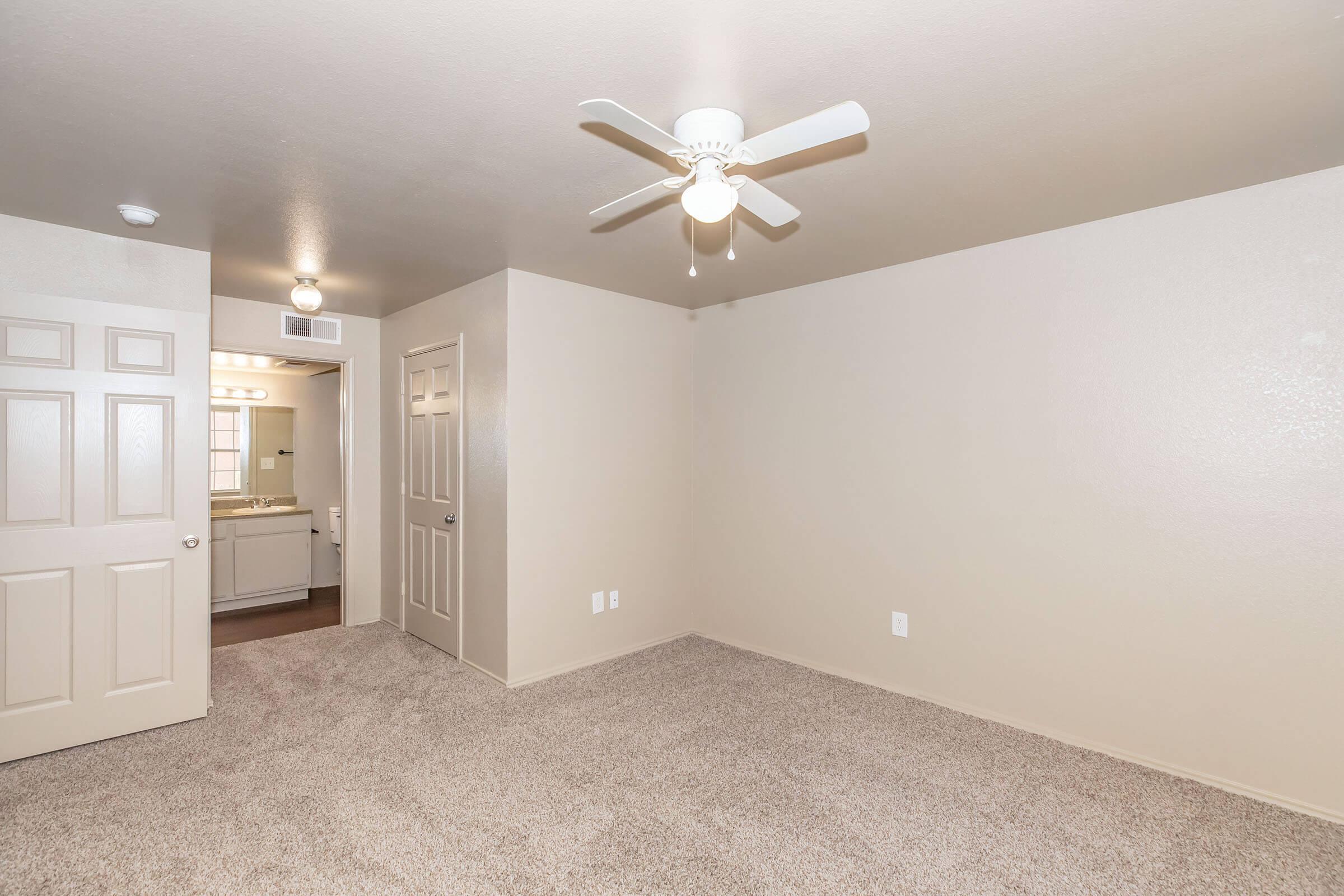 A spacious, empty room with light beige walls and carpeted flooring. A ceiling fan is visible, and a doorway leads to a bathroom area with bright lighting. The room has a calm and neutral aesthetic, suitable for various furniture arrangements.
