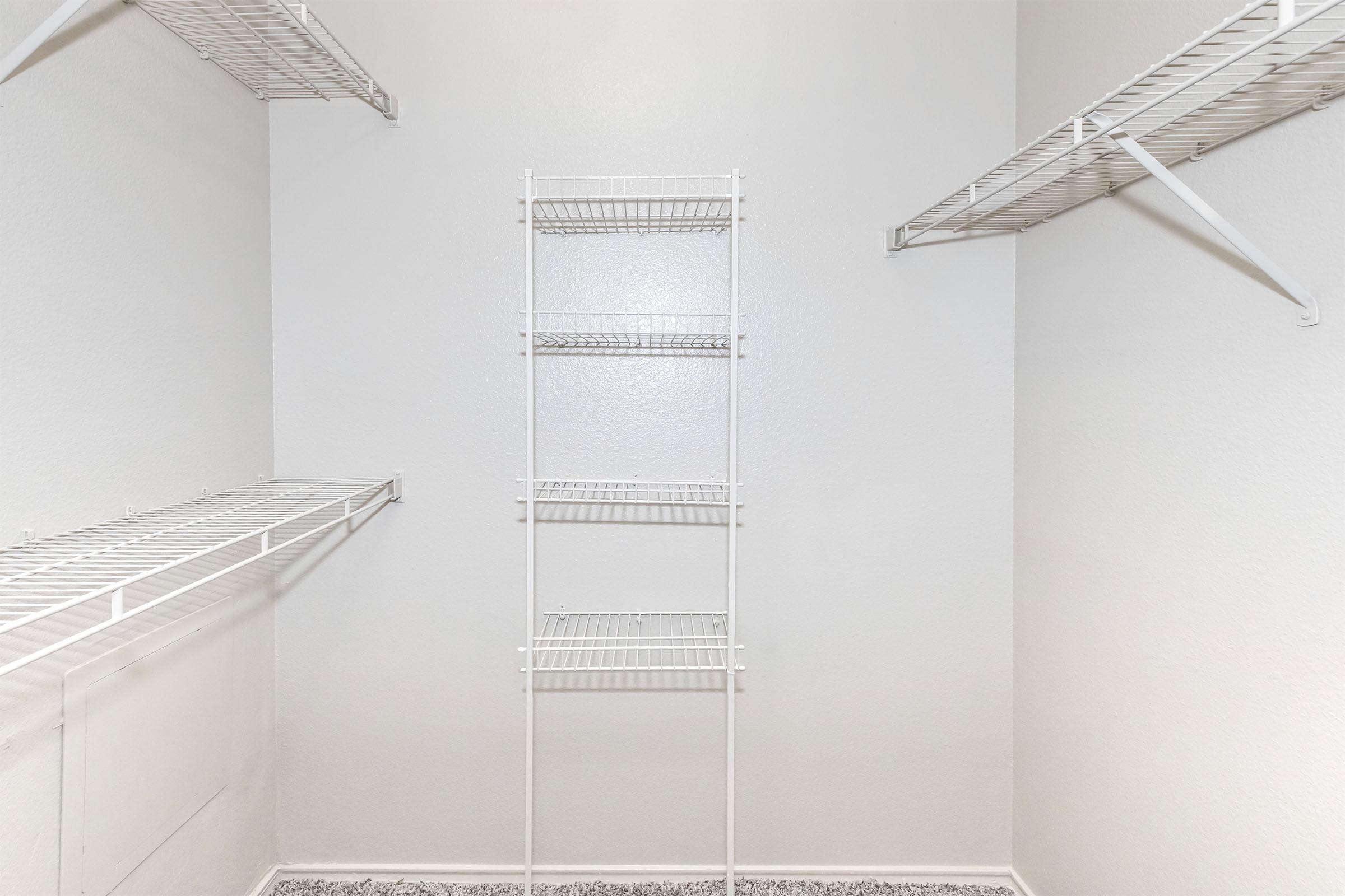 Empty closet with light gray walls. It features white wire shelving on both sides and a tall white wire rack in the center. The floor is covered with a textured surface. The overall appearance is clean and minimalistic, ideal for organization.