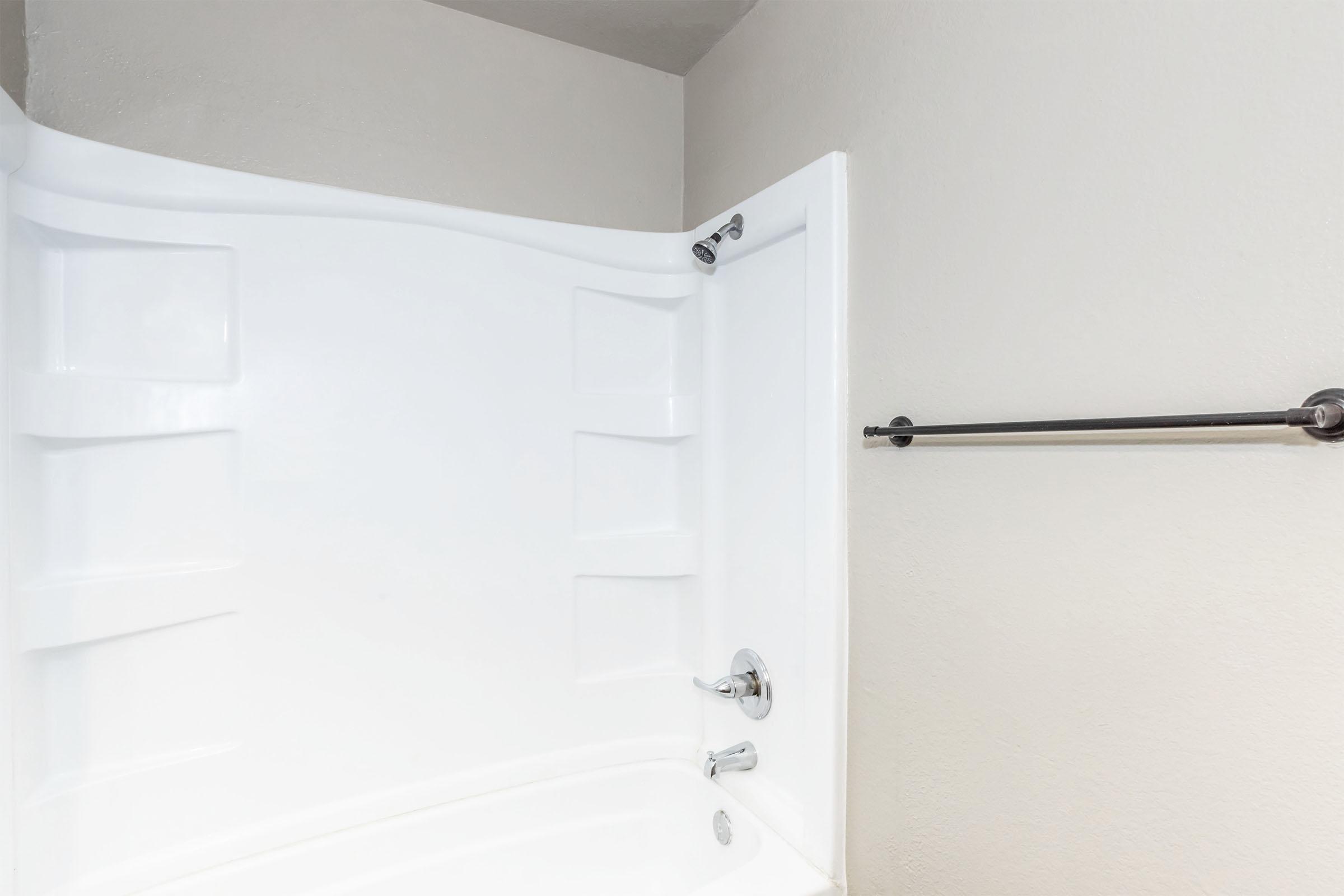A clean, modern bathroom featuring a white bathtub with a curved shower wall. The wall has built-in shelves for toiletries, and there's a towel bar mounted on the adjacent wall. The overall color scheme is light and neutral, creating a spacious and bright atmosphere.