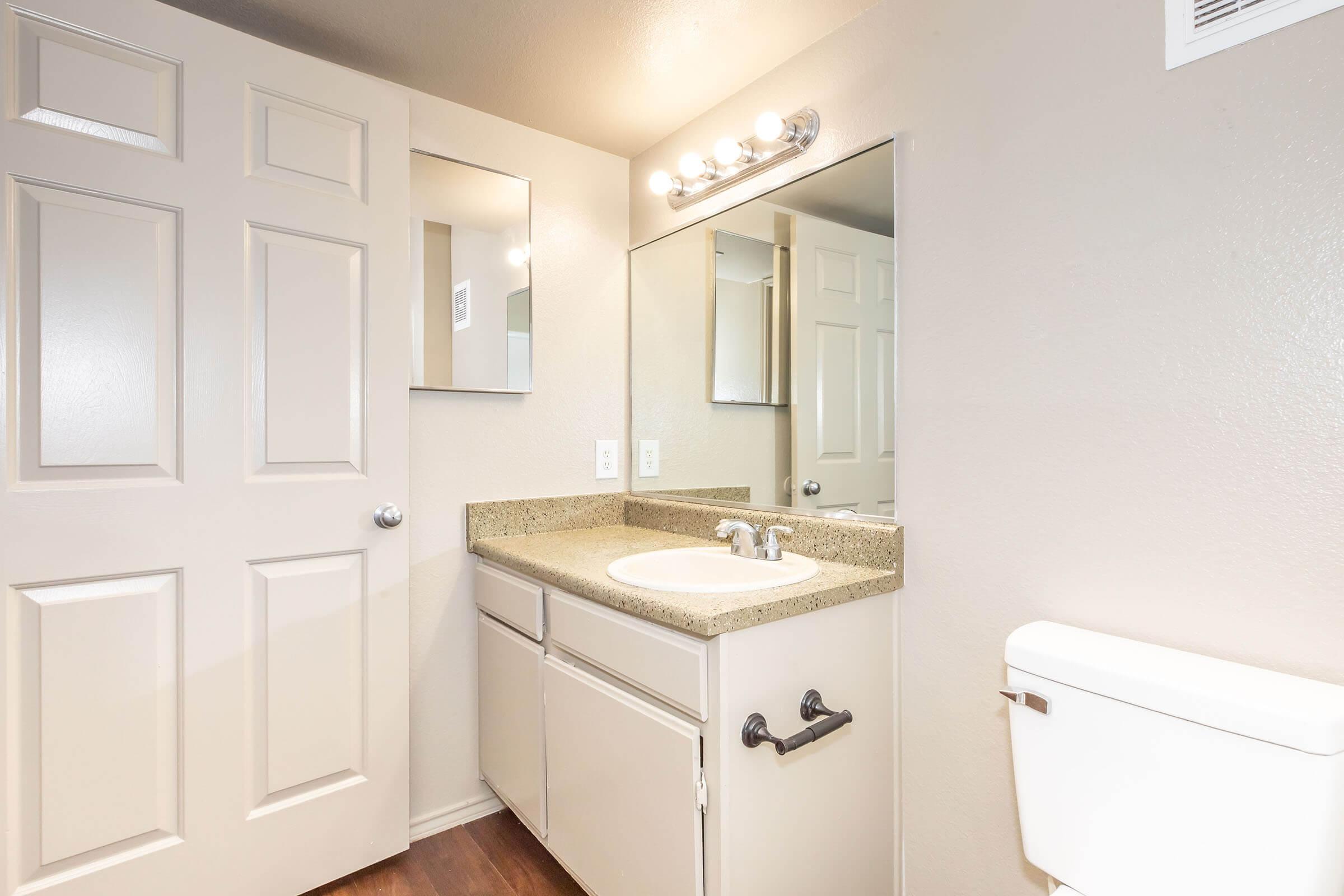 A clean, modern bathroom featuring a light-colored granite countertop with a sink, a large mirror above the sink, and a light fixture. The room has light gray walls, a wooden floor, a closed toilet, and a beige cabinet below the sink. A door with paneling is visible, adding depth to the space.