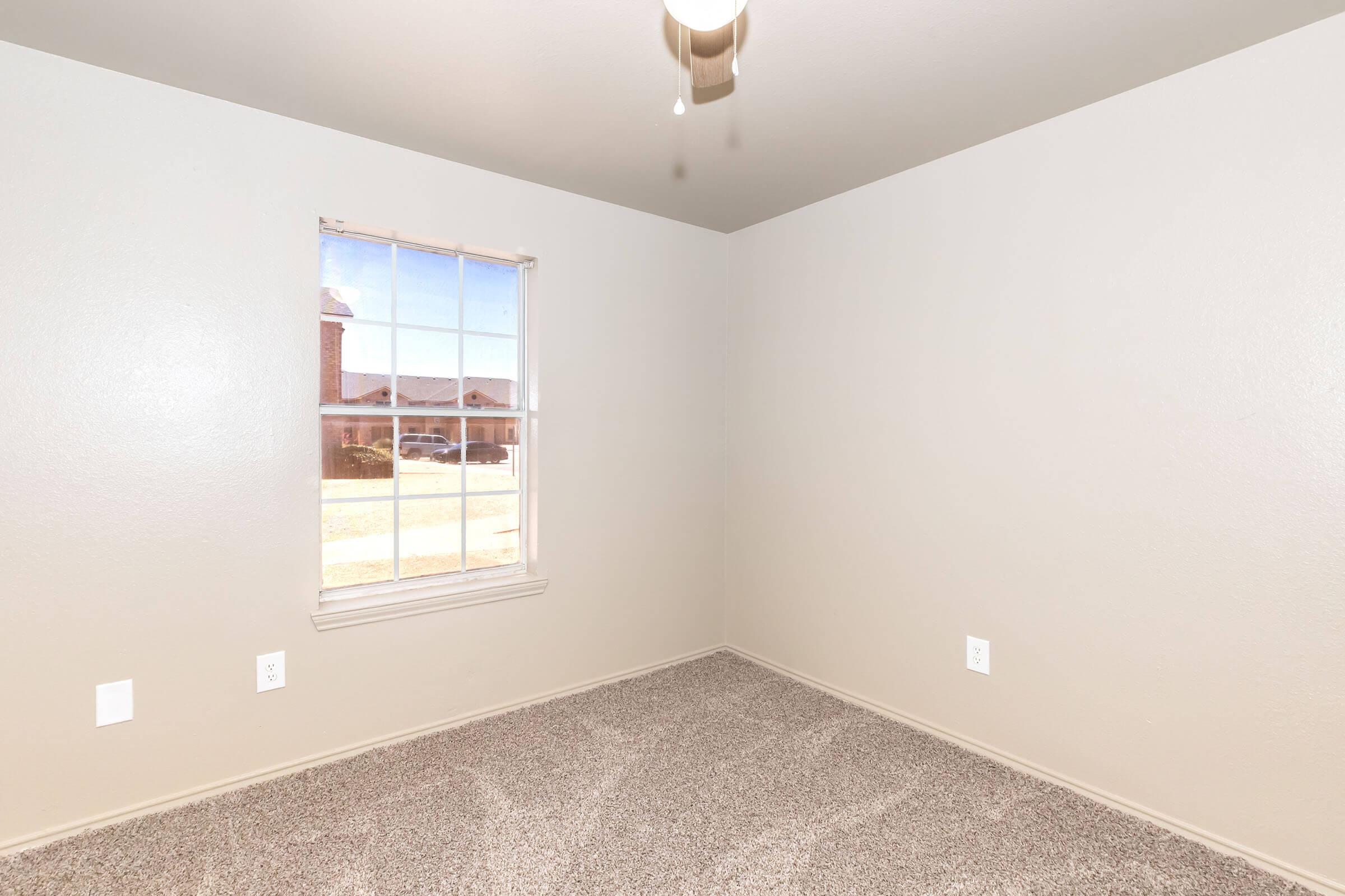 Empty room with light gray walls and carpeted floor. A window on the left allows natural light to enter, showing a glimpse of the outdoors. Ceiling fan hangs above, and there are electrical outlets on two walls. The space appears spacious and ready for furnishing.