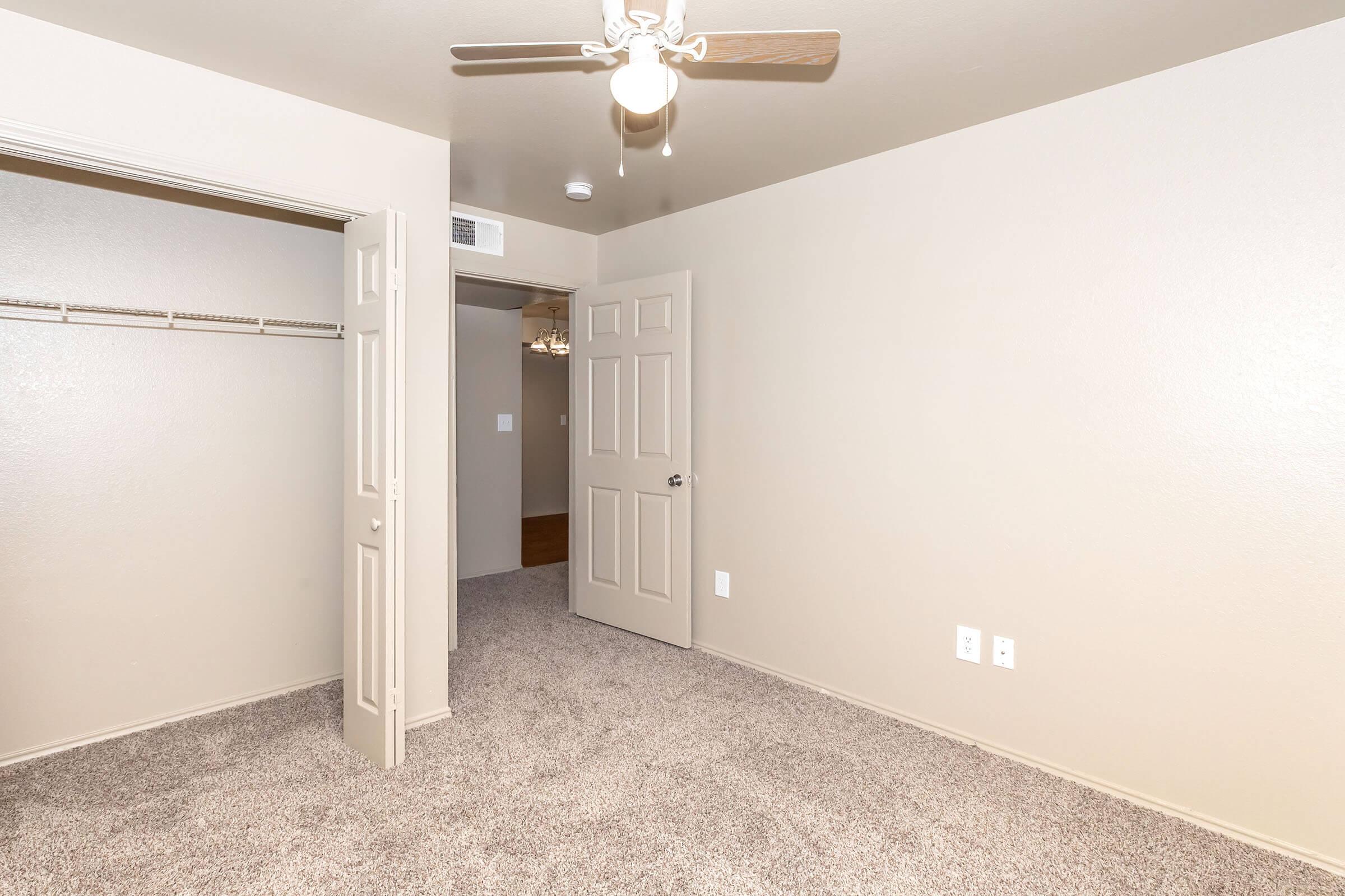 A spacious, neutral-colored room with light-colored walls and plush carpet. It features a ceiling fan, a door leading to another area, and a closet with shelving. Natural light comes from an unseen source, giving the room a bright appearance. The overall decor is simple and clean.