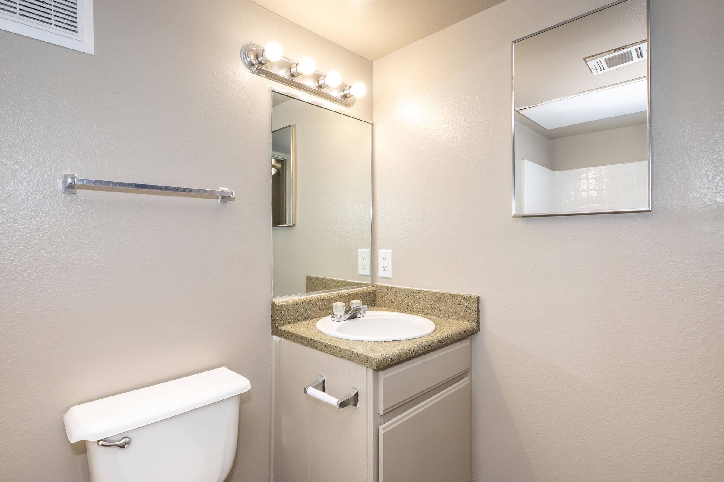 A clean bathroom featuring a white sink with a faucet on a granite countertop, a rectangular mirror above the sink, a wall-mounted light fixture with multiple bulbs, a towel rack, and a white toilet. The walls are painted in a light, neutral color, creating a bright and tidy space.