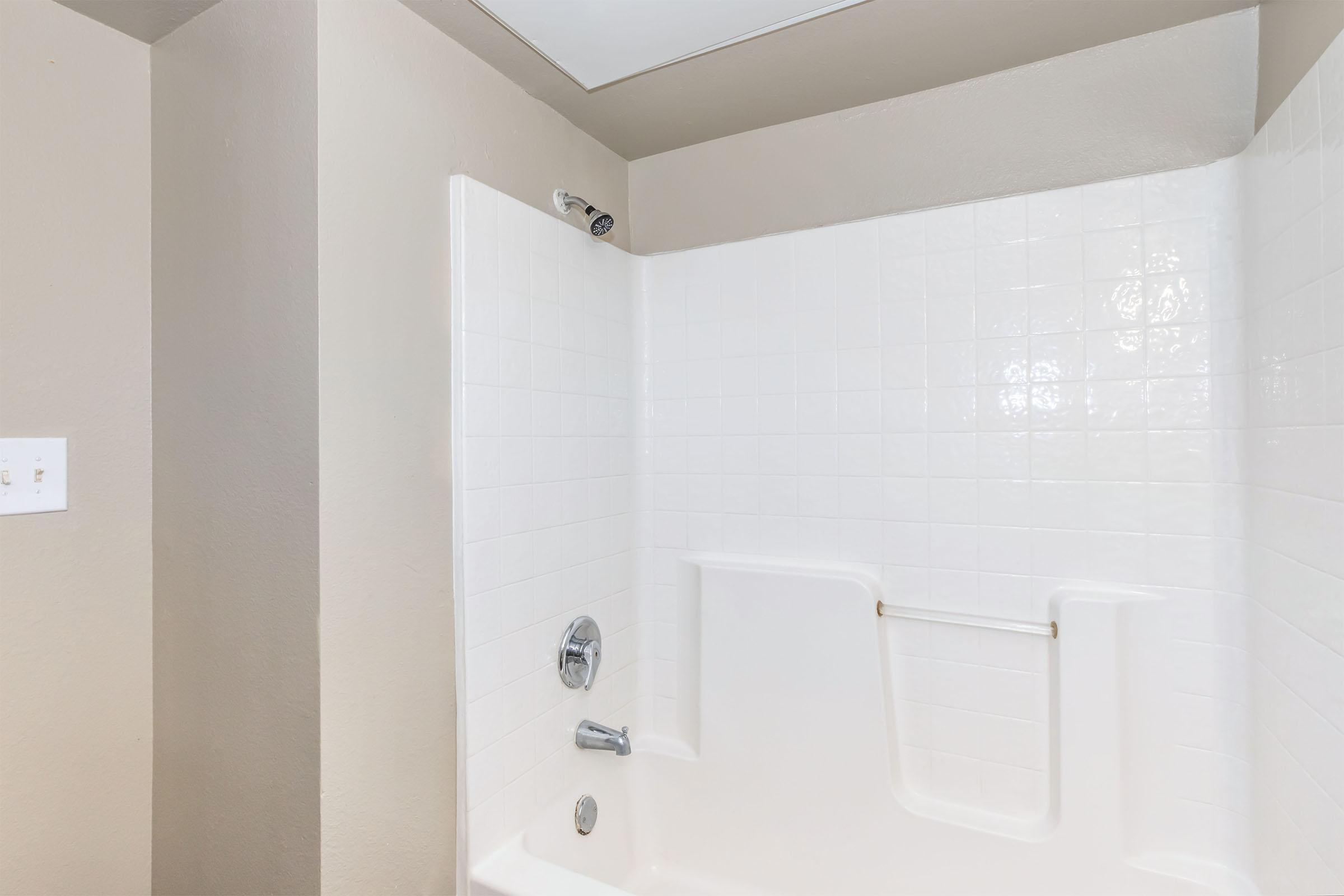 A clean bathroom shower area features a white tile wall, a white bathtub with a low wall, and stainless steel fixtures. The walls are painted in a neutral color, creating a bright and open space. Natural light illuminates the area through a small overhead window.
