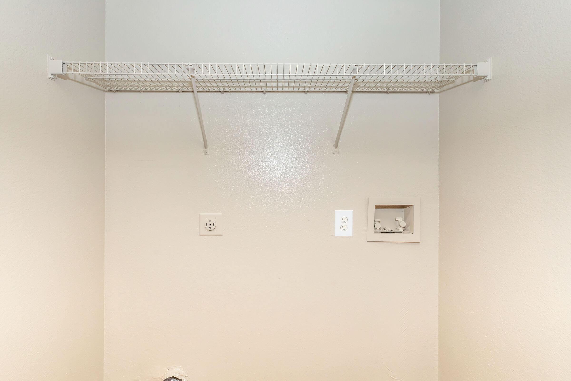 Empty closet space with a white wire shelf mounted on the wall, two electrical outlets on the right side, and a small recessed box below the shelf. The walls are painted a light color, creating a clean and minimalistic look.