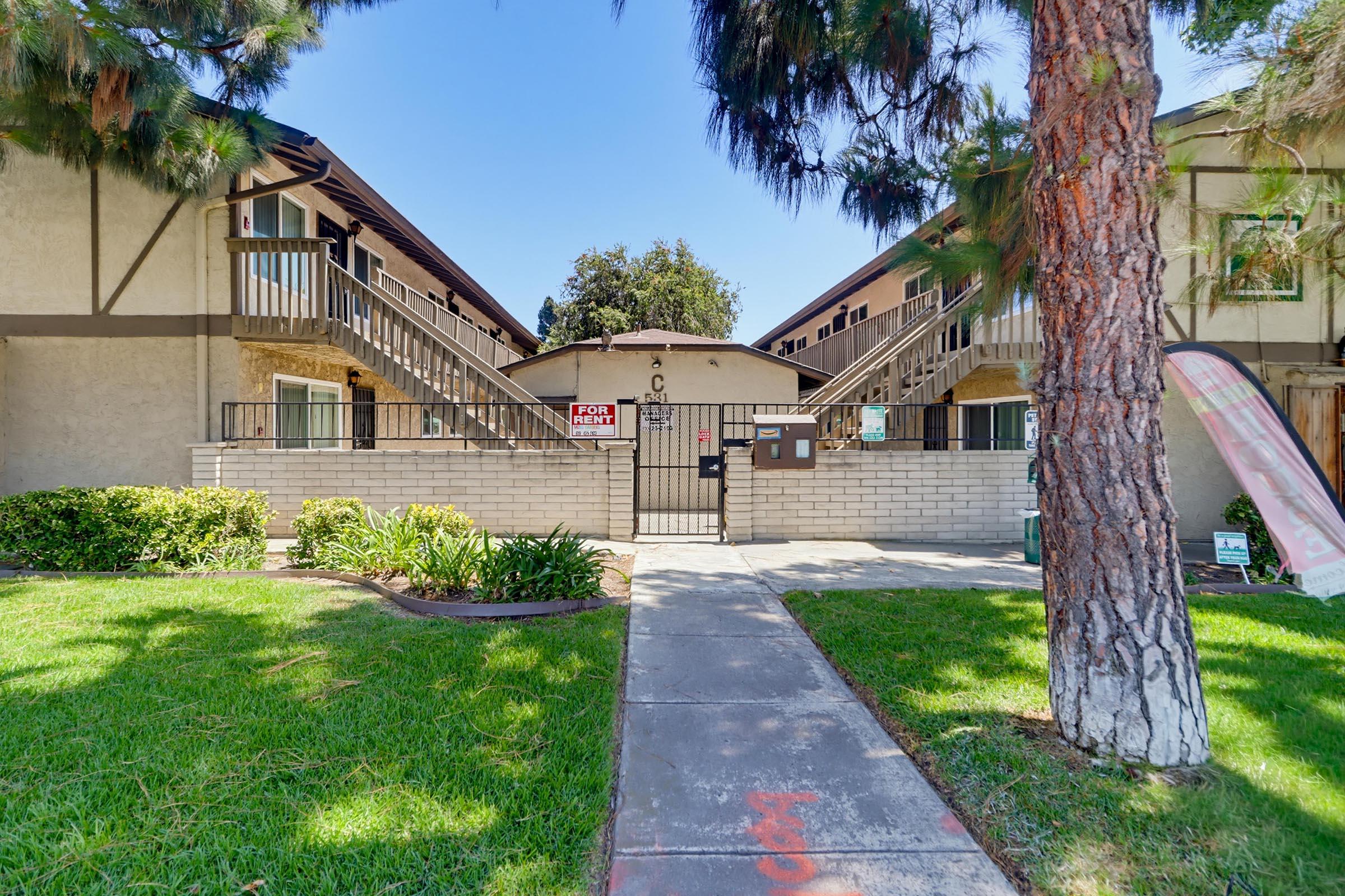 A two-story apartment complex with a central entrance and staircases leading to upper units. Grass and shrubs are maintained in the front yard, and a "For Rent" sign is visible near the entrance. The building is framed by tall trees, providing some shade in the area.