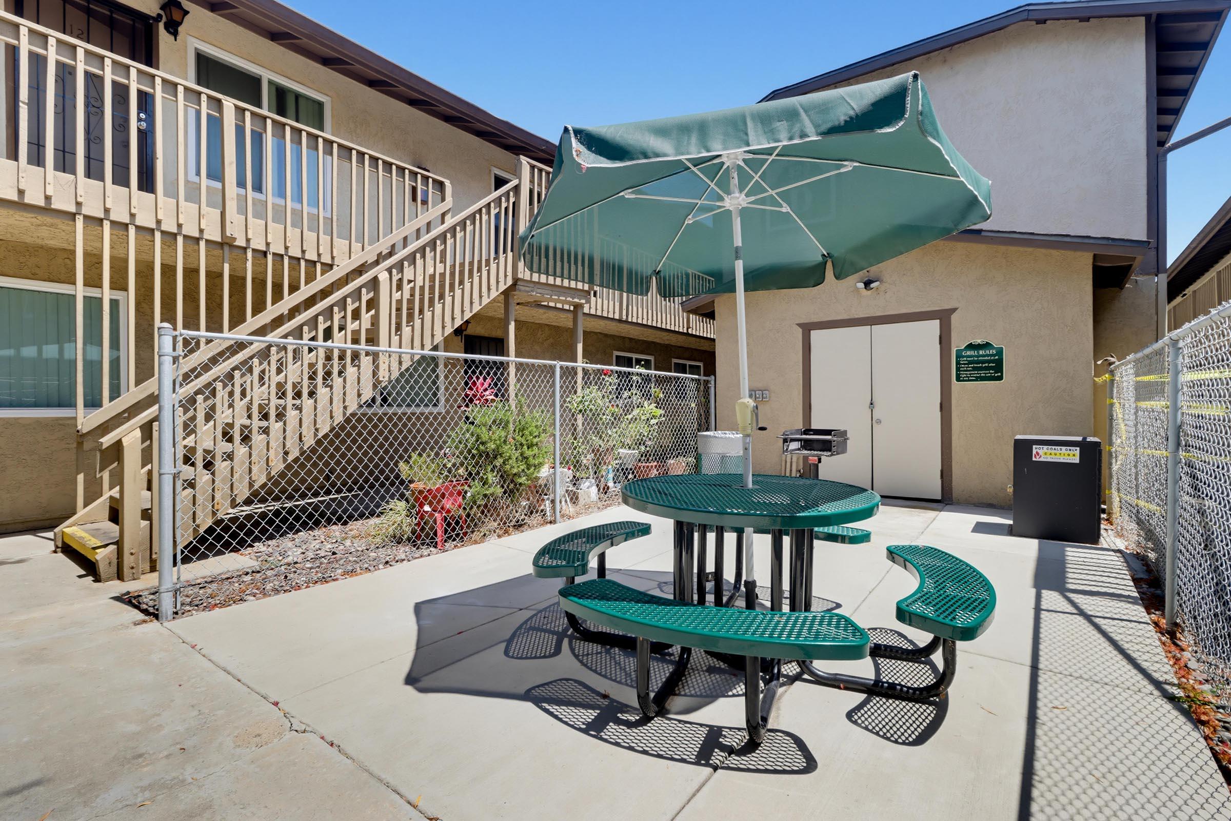 A small outdoor patio area featuring a green picnic table with benches, a large umbrella for shade, and a barbecue grill nearby. The setting includes a two-story building in the background and a fenced area with plants.