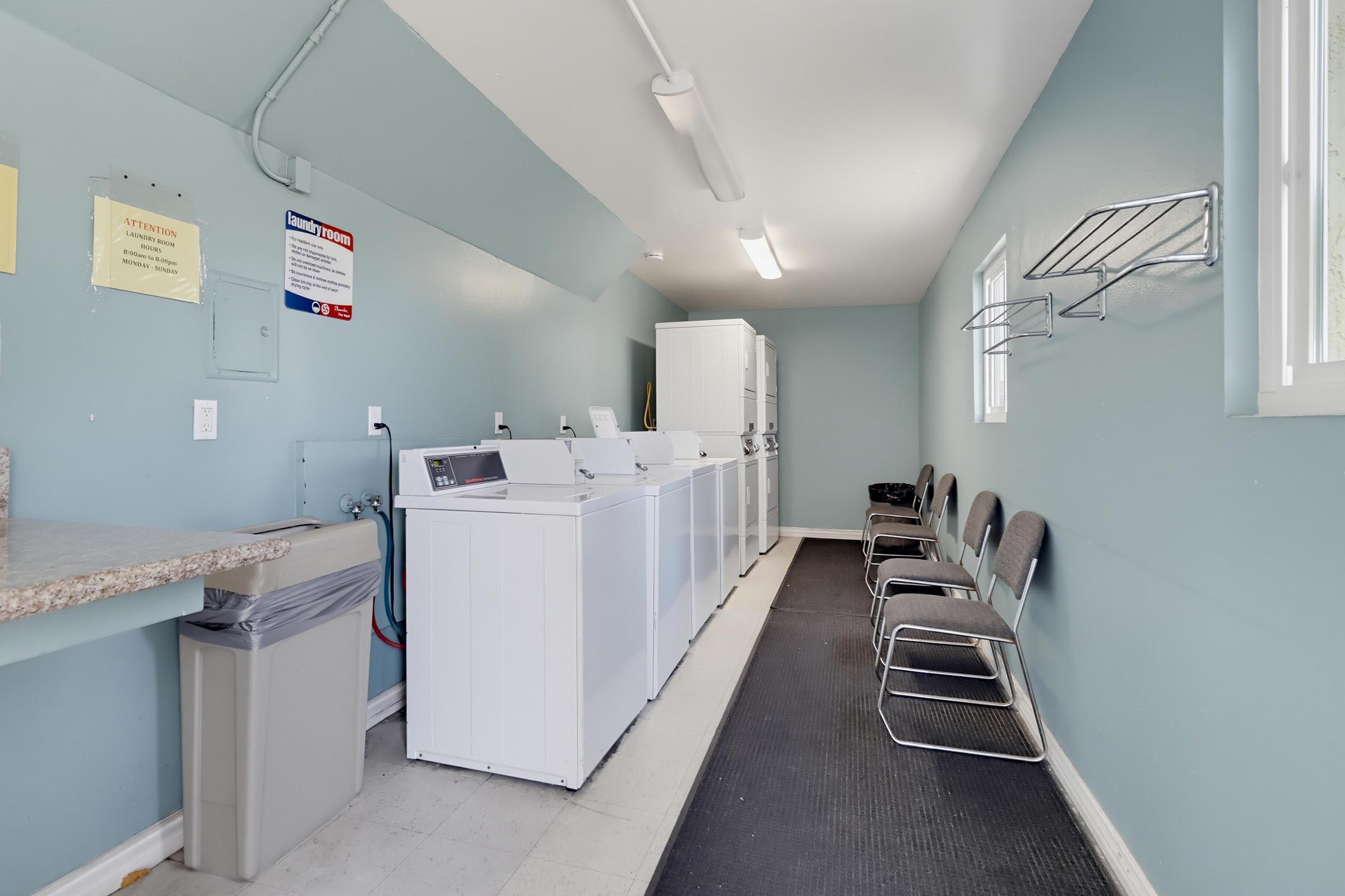 A laundry room featuring several white washing machines along one wall, a countertop with a trash can, and a row of metal chairs. The walls are painted light blue, and there are notices posted on the walls. A black mat is positioned on the floor for comfort. Natural light comes through a window.