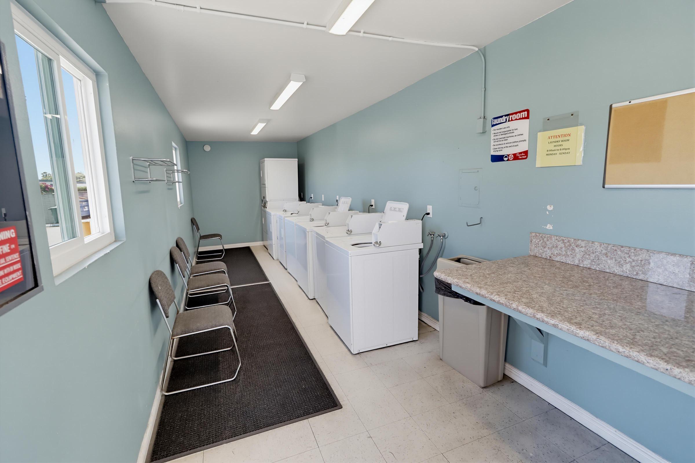 A clean and bright laundry room featuring several washing machines aligned against one wall. There are chairs along the opposite wall, a countertop for folding clothes, and a trash bin. The walls are painted light blue, and there are windows providing natural light.