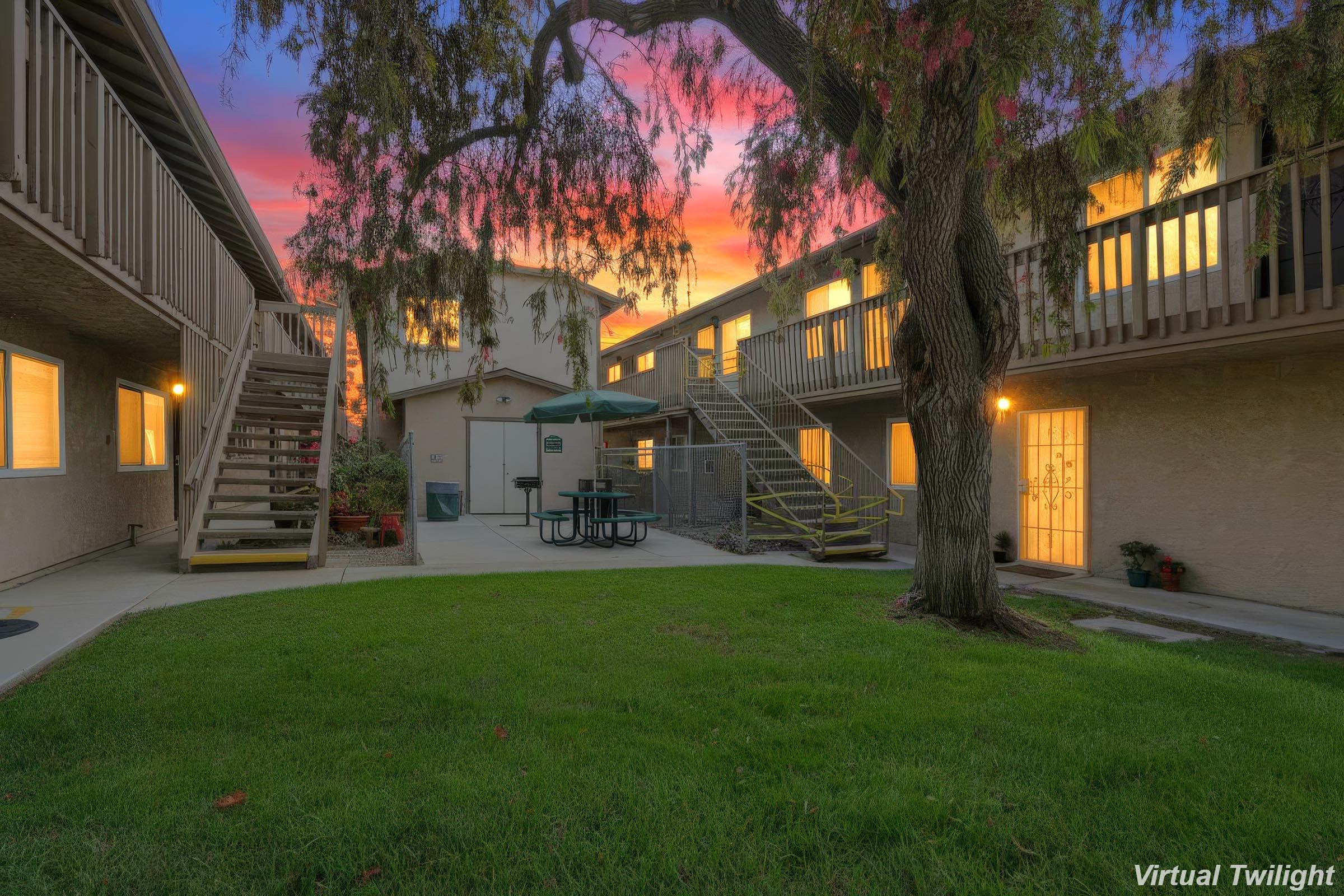 A serene courtyard scene at dusk, featuring grassy areas, a picnic table surrounded by trees, and buildings with illuminated windows. The sky is vibrant with shades of orange and purple as the sun sets, creating a peaceful atmosphere in the residential complex.