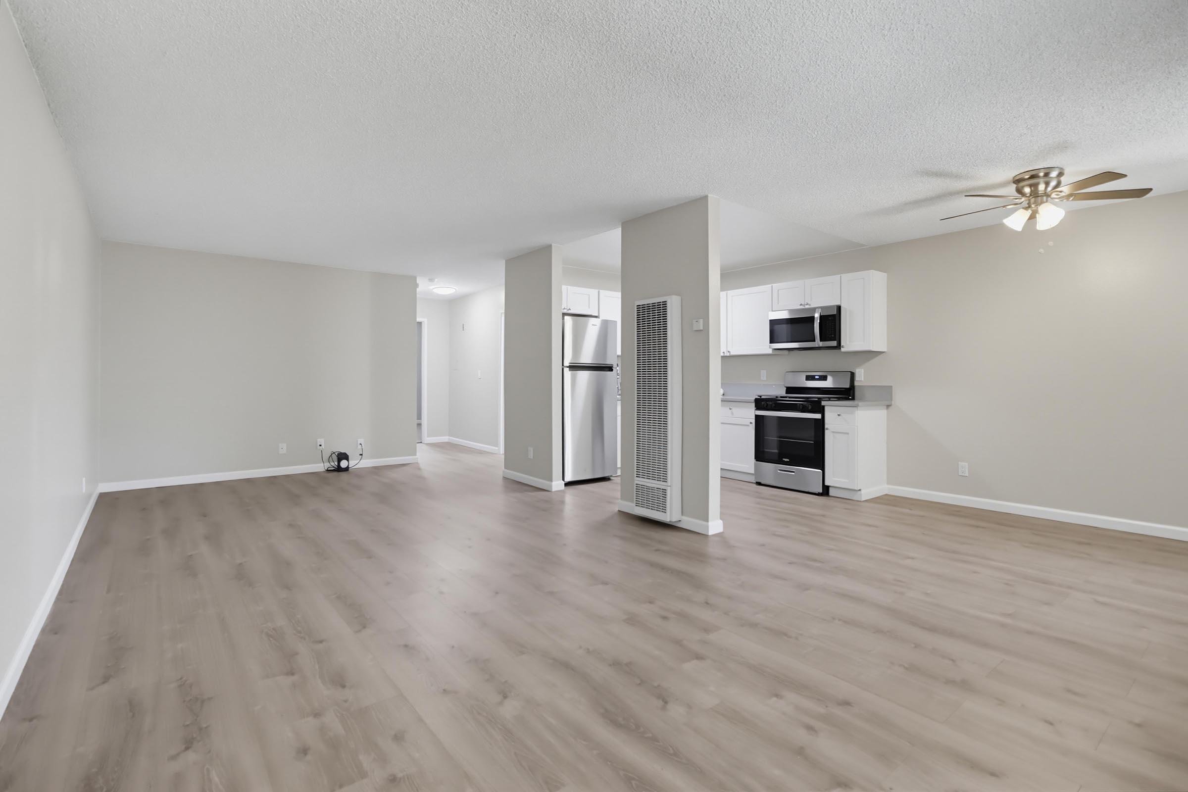 Spacious living area featuring light-colored walls and a ceiling fan. The open layout leads to a kitchen with modern appliances, including a stainless-steel refrigerator and oven. The floor is finished with light wood laminate, creating a bright and inviting atmosphere.