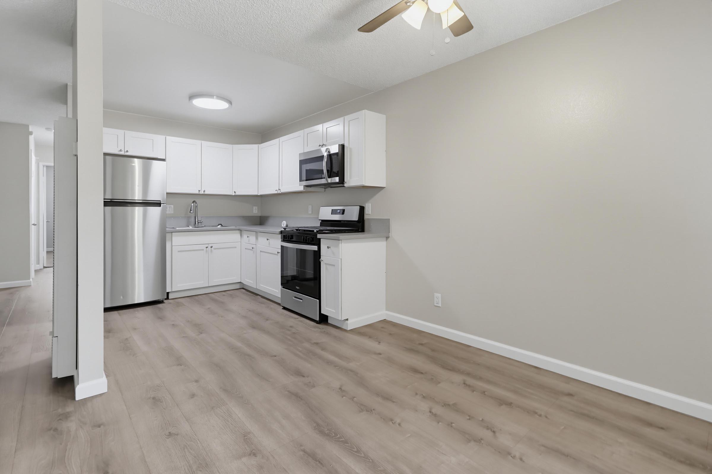 Modern kitchen featuring white cabinetry, stainless steel appliances including a refrigerator and stove, and a microwave. The space has light-colored walls, a ceiling fan, and wooden floors, creating a bright and open atmosphere. Ideal for a small apartment or studio layout.